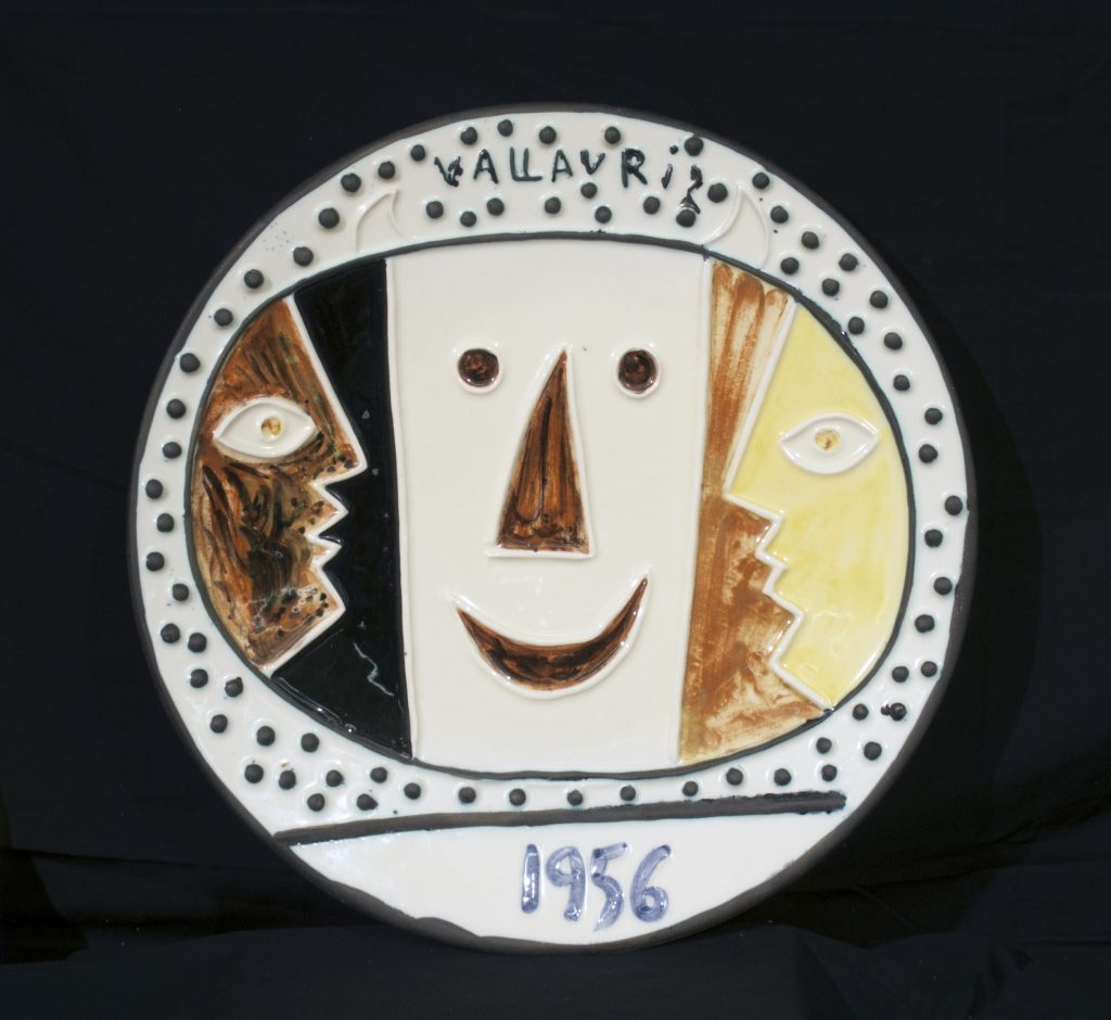 A photo of a ceramic plate by Pablo Picasso. The plate features the side profile of two faces flanking the front of a third face. The date 1956 is inscribed on the bottom of the plate in blue.