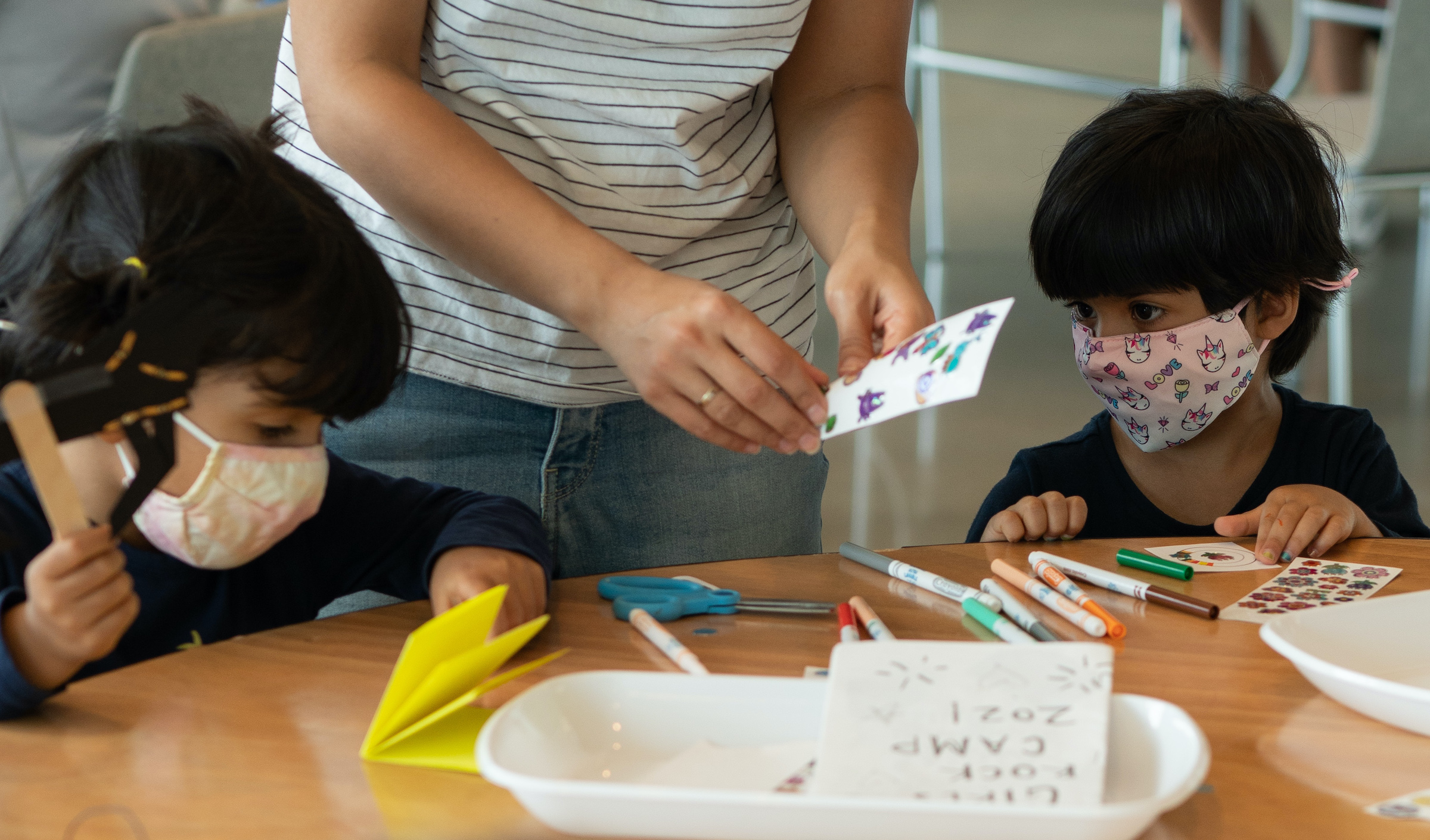 Two children sit at a table with a parent, making signs with various materials spread around them.