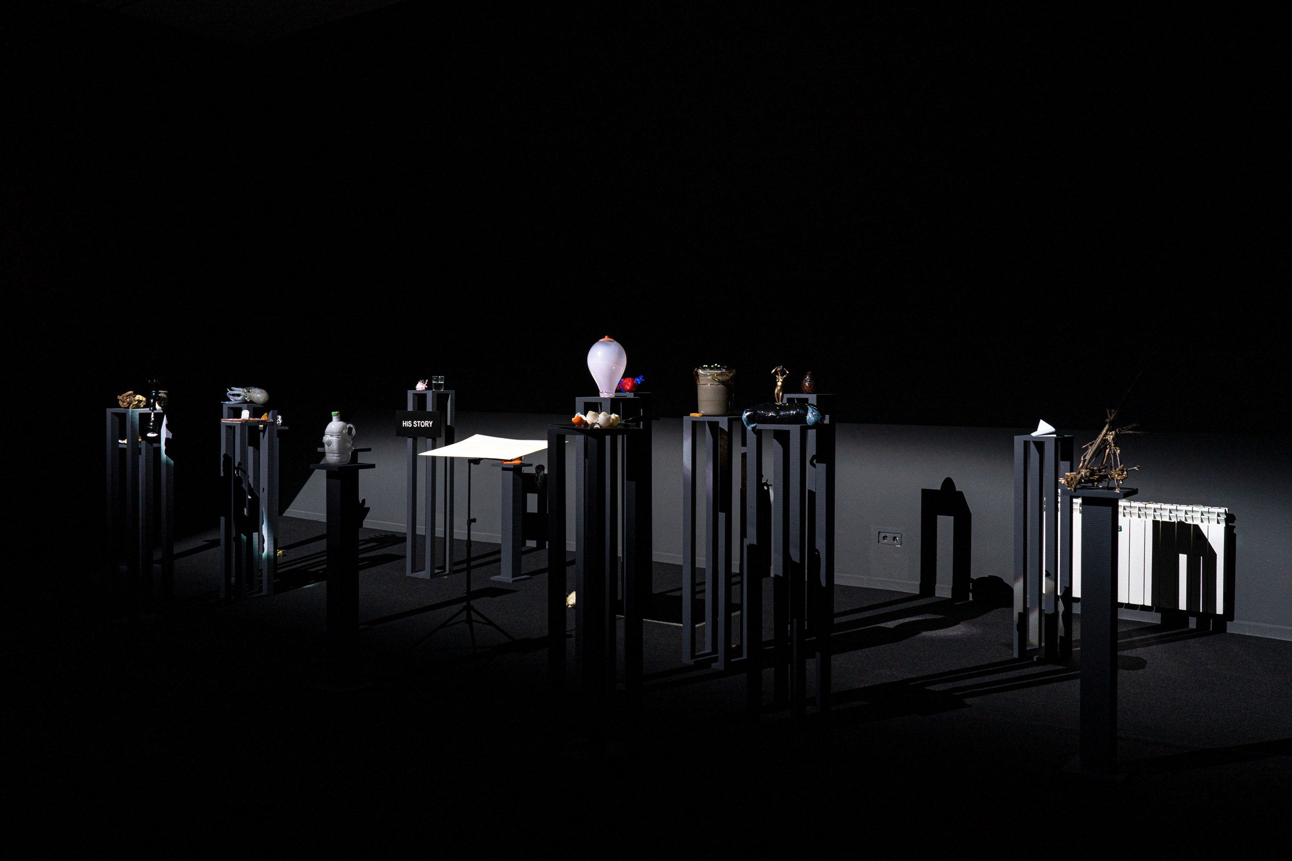 An artwork by French artist Laure Prouvost is made up of numerous black platforms, with a variety of objects including glass sculptures, placed on top. The entire room is black with spotlights illuminating the installation.