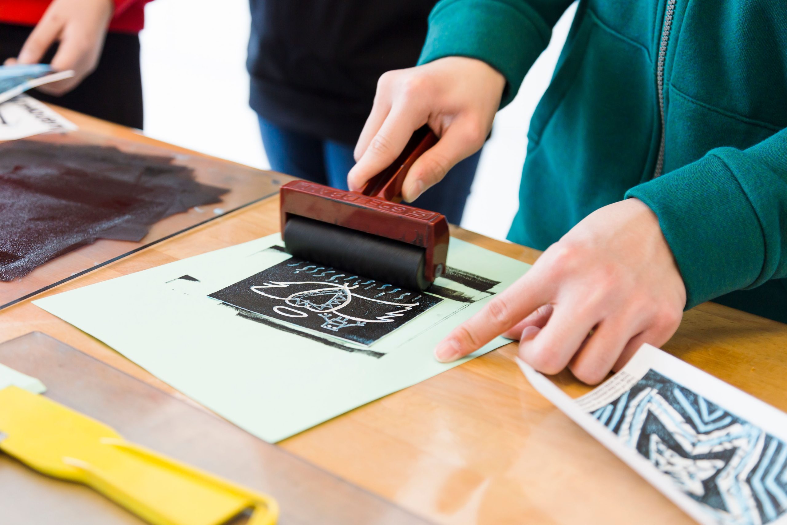 A photograph shows the hands of a person rolling ink onto a linocut print.