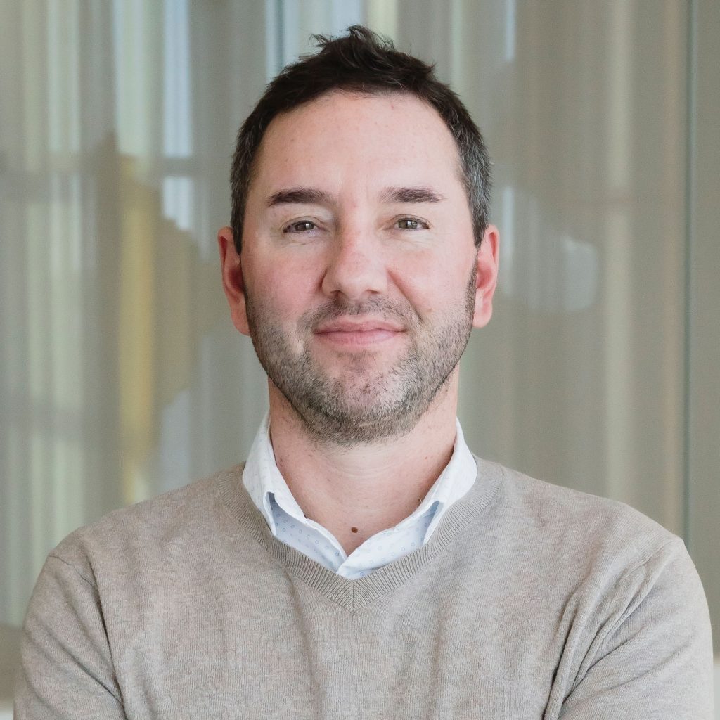 Head-and-shoulders portrait of a person with short dark hair and light stubble, wearing a light sweater over a collared shirt, standing in front of softly lit curtains.