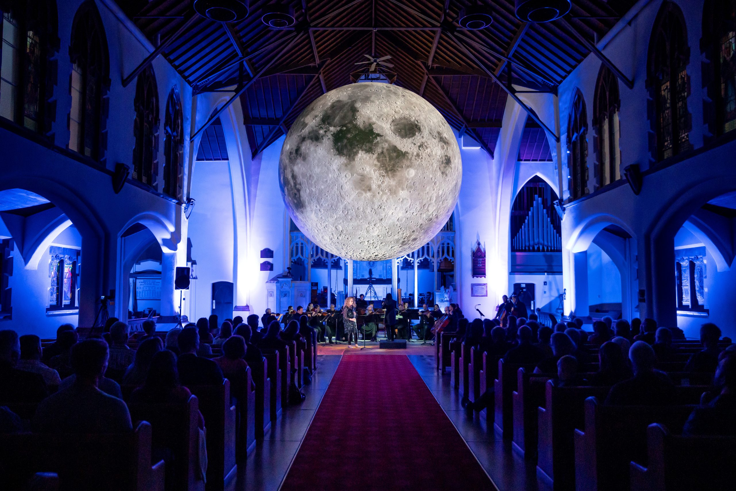 "An indoor event with a large, detailed replica of the moon hanging from the ceiling of a church, illuminated by blue lighting. An audience sits in pews, facing a stage where musicians are performing."