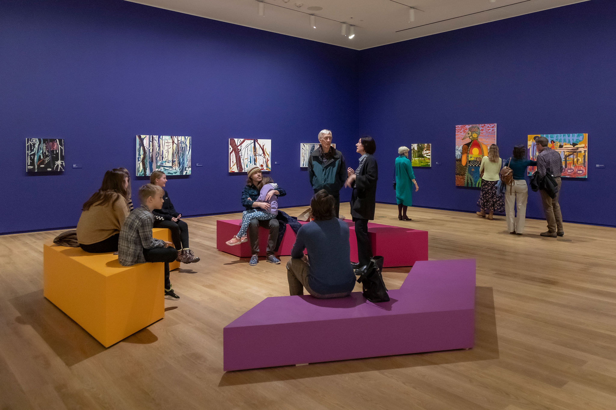 A group of people are gathered in a modern art gallery with vibrant purple walls. Several colorful paintings are displayed on the walls, and visitors are engaged in observing the artwork, conversing, or sitting on geometric benches in shades of yellow, pink, and purple.