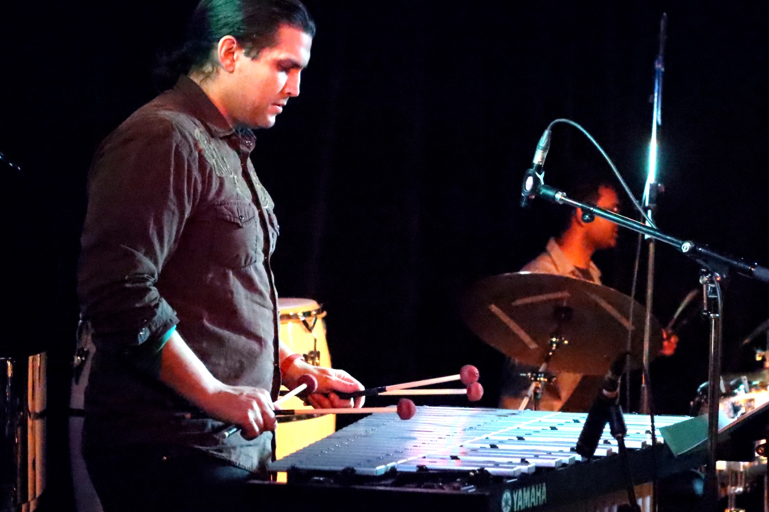 A musician playing a Yamaha vibraphone on stage, holding four mallets. He is wearing a dark button-up shirt and has his hair tied back. In the background, another musician is playing the drums, partially obscured by a microphone stand. The stage is dimly lit with a dark backdrop.
