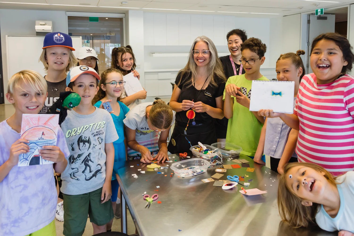 Group of smiling children holding up pieces of art in a studio as two staff members look on, happy.