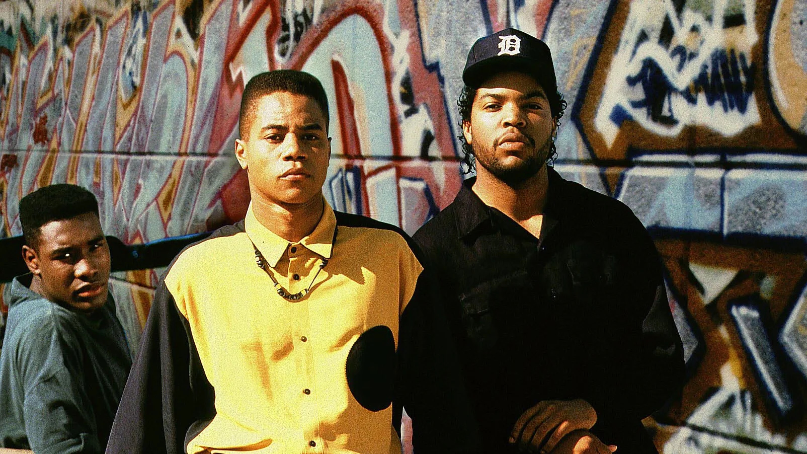 Three young men stand in front of a graffiti-covered wall, staring seriously at the camera. One wears a yellow button-up shirt with black accents and a beaded necklace, another wears a black outfit with a Detroit Tigers baseball cap, and the third, slightly behind them, wears a dark blue shirt. The image is vibrant and sunlit, evoking a gritty, urban atmosphere with a strong sense of identity and presence.