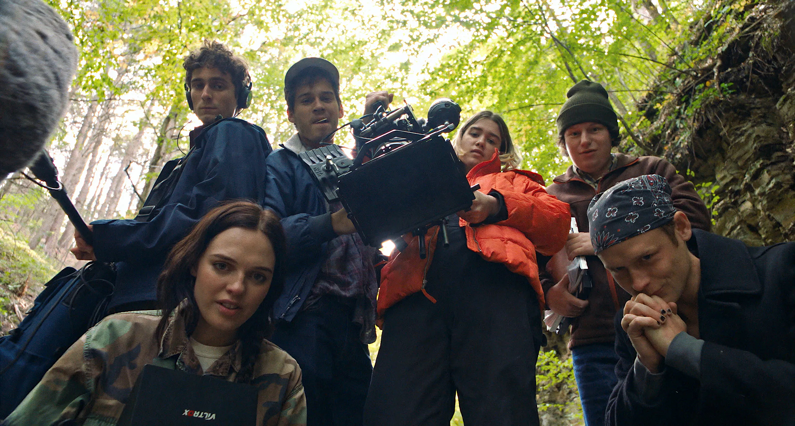Six young adults gather in a lush, green forest, all looking down toward the camera from above. One person holds a professional video camera, another wears headphones while gripping a long boom microphone, and others carry notebooks or gear. They’re dressed in casual outdoor clothing—some in jackets or beanies—and stand closely together, as sunlight filters through the leaves above.