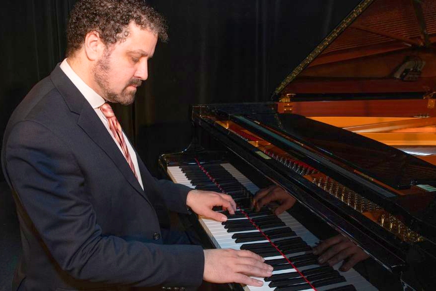 A pianist in a dark suit and striped tie is playing a grand piano on stage. He has curly hair and a beard, and he is focused on the keys as he performs. The piano’s glossy surface reflects his hands and the surrounding stage lighting. The background is dimly lit with a black curtain.