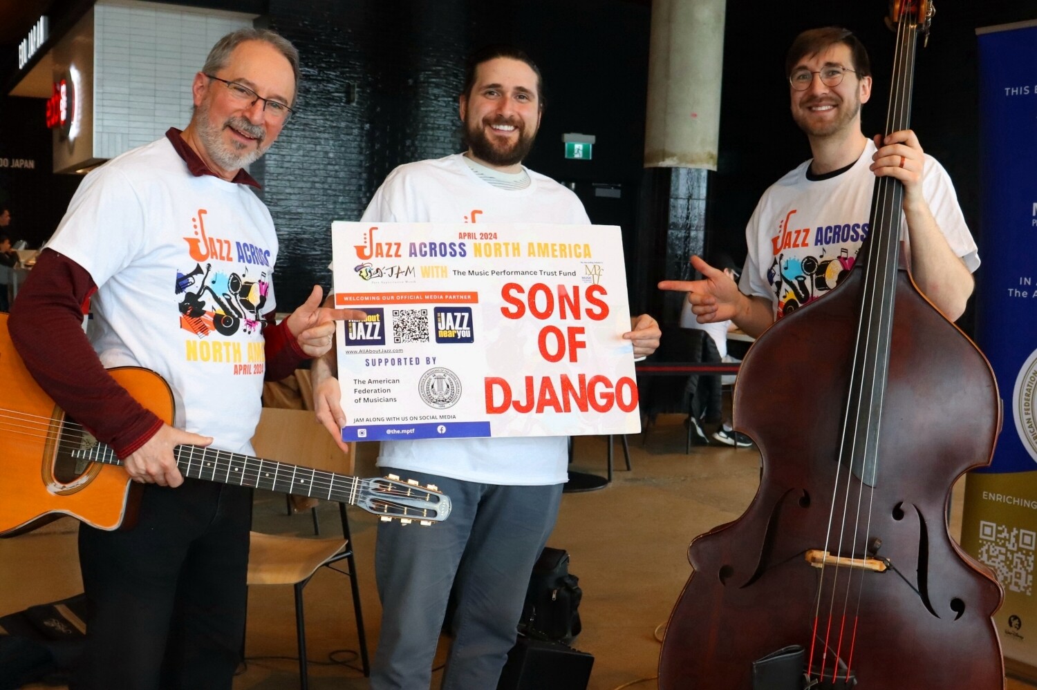 Three musicians from the group “Sons of Django” pose together at a jazz event, wearing matching “Jazz Across North America” t-shirts. One musician holds an acoustic guitar, another holds a double bass, and the third holds a sign with the band’s name. They are smiling and pointing at the sign, standing in a modern indoor venue with banners and chairs in the background.
