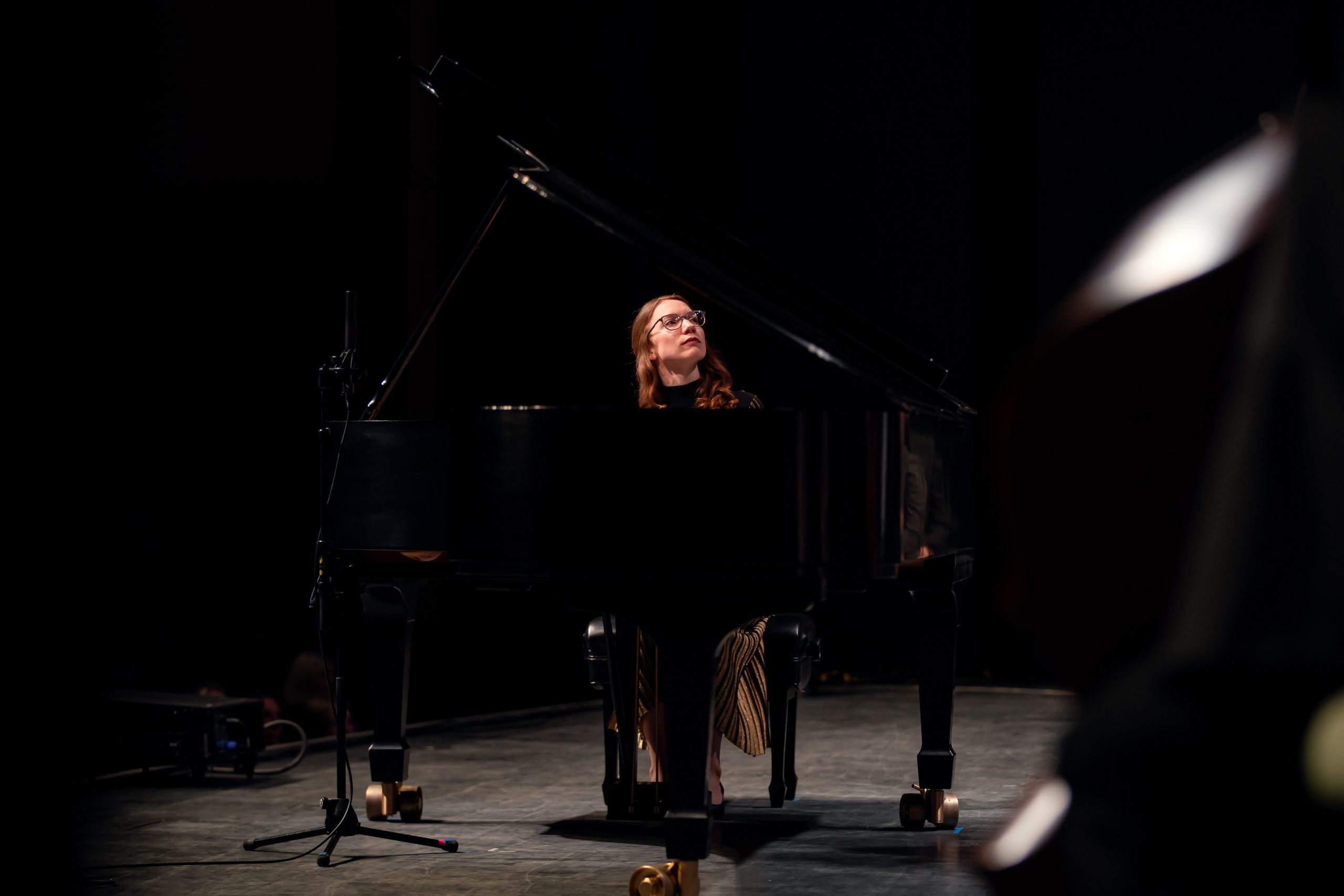 A woman with long hair and glasses sits at a grand piano on a dimly lit stage. She looks upward thoughtfully as she plays. A microphone stand is positioned beside the piano, and a faint silhouette of another instrument appears in the foreground.
