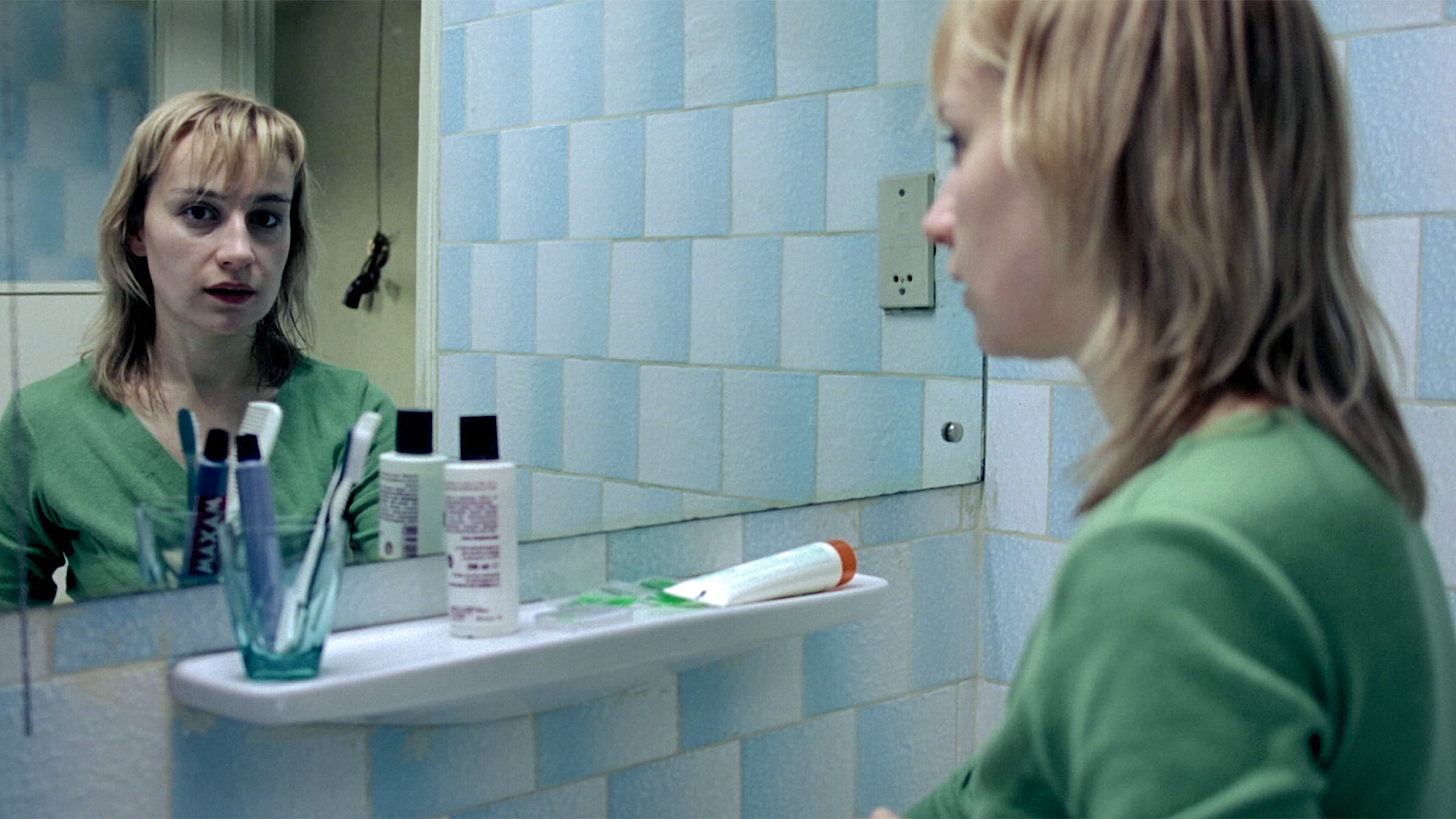 A woman with shoulder-length blonde hair and a green top stands in a bathroom, looking at herself in the mirror with a tense, pensive expression. The bathroom walls are covered in blue and white tiles. A white shelf below the mirror holds several toothbrushes in a glass, two bottles, and a tube of toothpaste. The atmosphere is stark and somber.