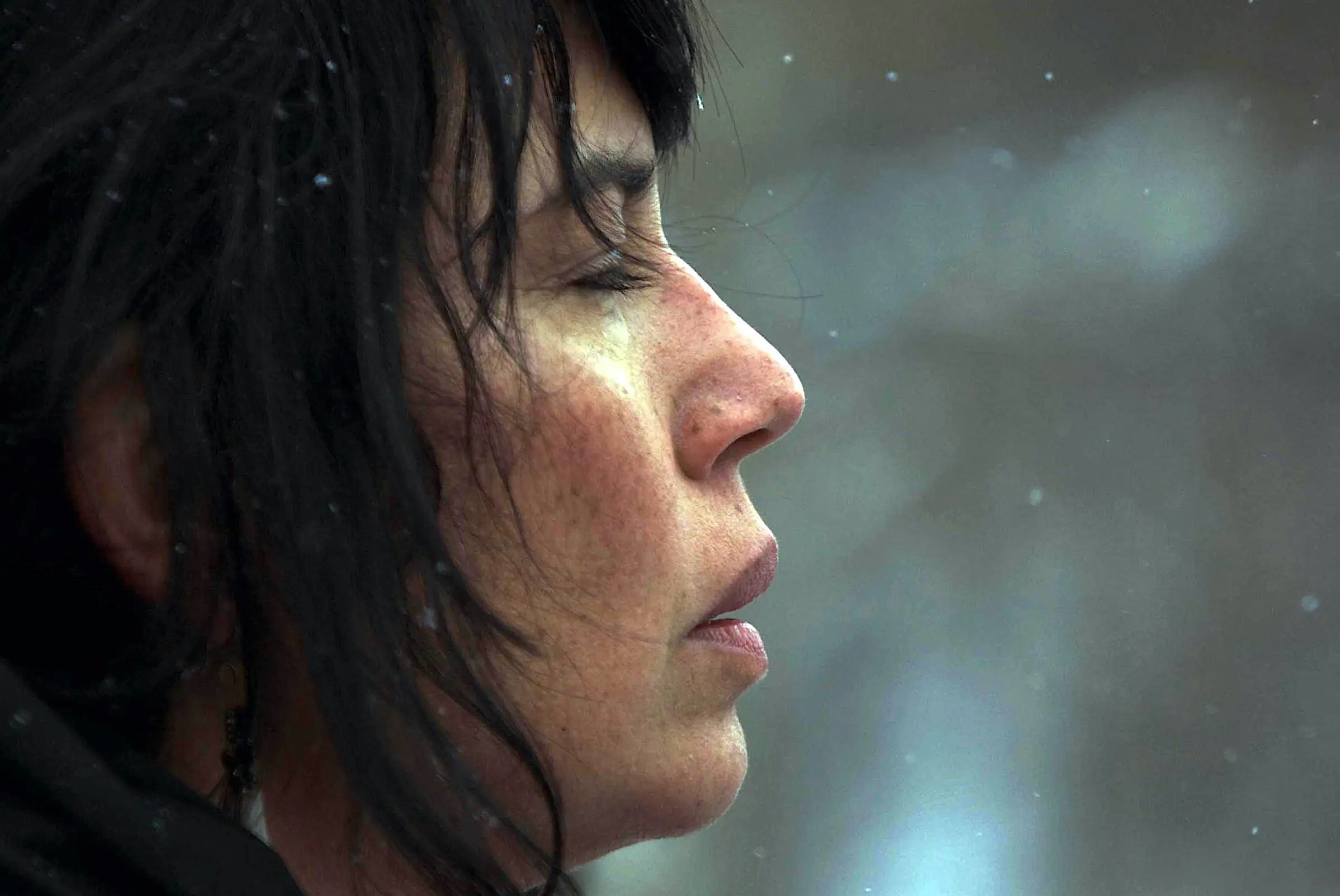 Close-up profile of a woman with closed eyes and long dark hair, her face softly lit against a blurred grey background. Snowflakes fall around her as she appears deep in thought or meditation.