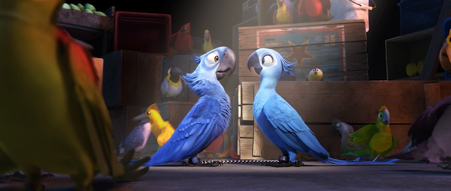 Two animated blue macaws stand side by side on the floor of a dimly lit room, chained together by their ankles. They look at each other with concerned expressions. Around them are colorful parrots and wooden crates, some stacked and some open, hinting at a smuggling operation. A spotlight shines down on the blue macaws, drawing attention to them in the crowded scene.