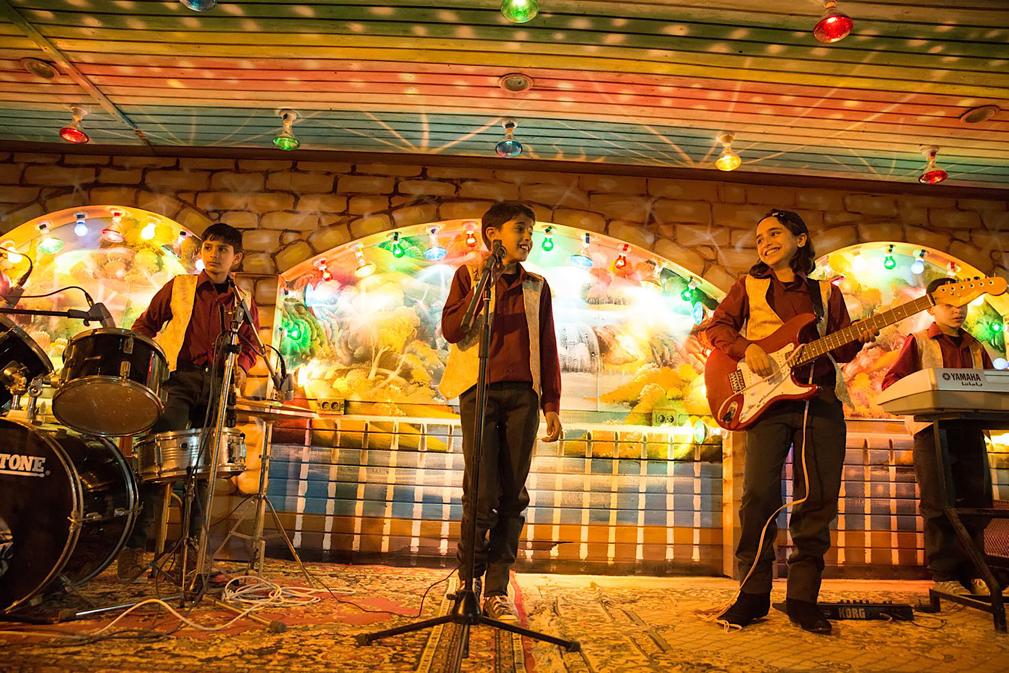 A group of children performs as a band on a brightly lit stage decorated with colourful lights and painted murals. A boy sings at the microphone, another plays drums, a girl smiles while holding an electric guitar, and another boy plays a keyboard. The atmosphere is warm and festive.