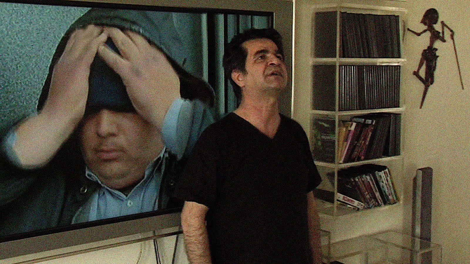 A man stands in a living room looking upward with a pained expression. Behind him, a large TV screen shows another man covering his face with his hands. Shelves filled with DVDs and books are visible to the side.