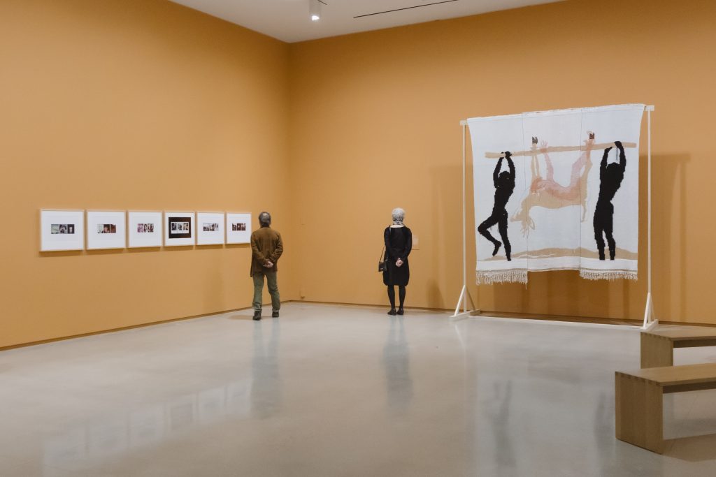 Two visitors stand in a gallery with orange walls, viewing framed photographs and a large textile artwork displayed on a freestanding frame.