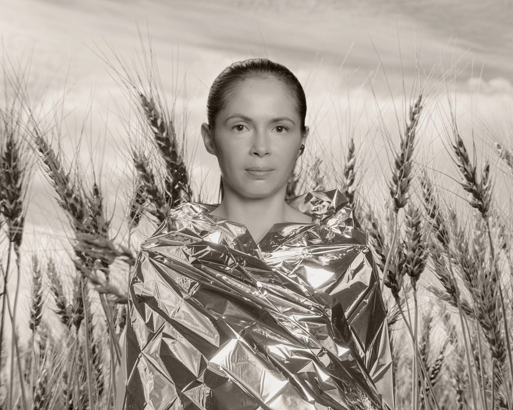 A person stands in a wheat field wearing a reflective metallic blanket that shimmers in the light. The portrait is in black and white, with tall wheat stalks surrounding the person under a softly clouded sky. The overall mood feels calm and contemplative, blending natural and futuristic elements.