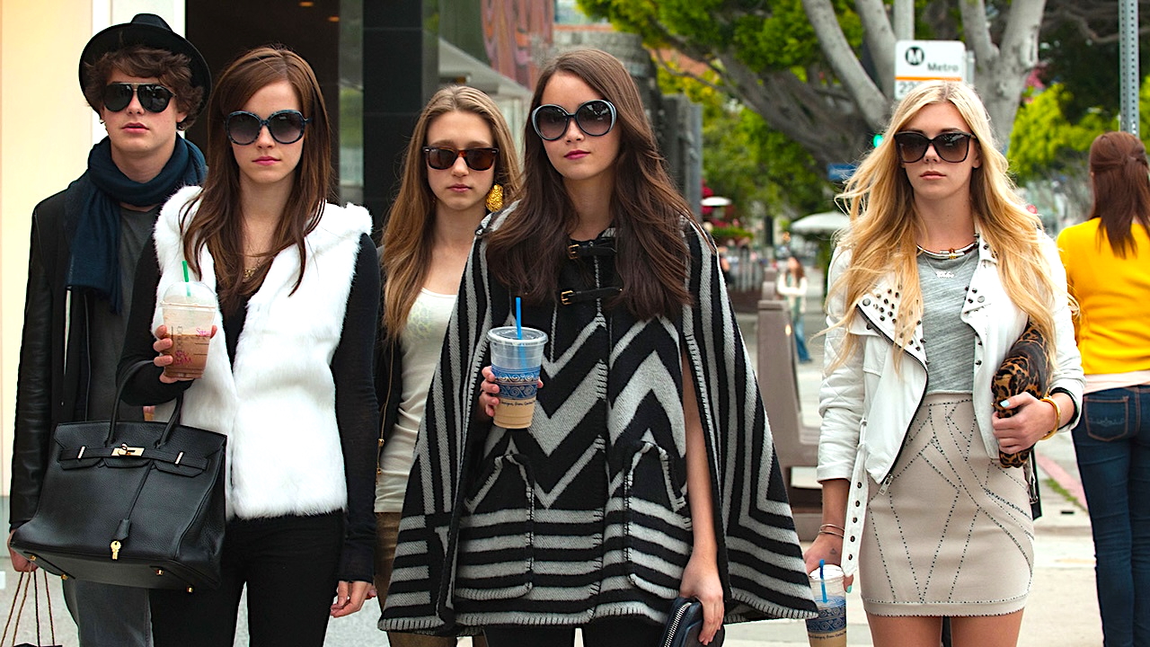 A stylish group of five young adults walk confidently down a city sidewalk, all wearing oversized sunglasses and fashionable outfits. Two carry iced drinks, and one holds a large black handbag. Trees, pedestrians, and storefronts line the background, evoking an upscale urban setting.