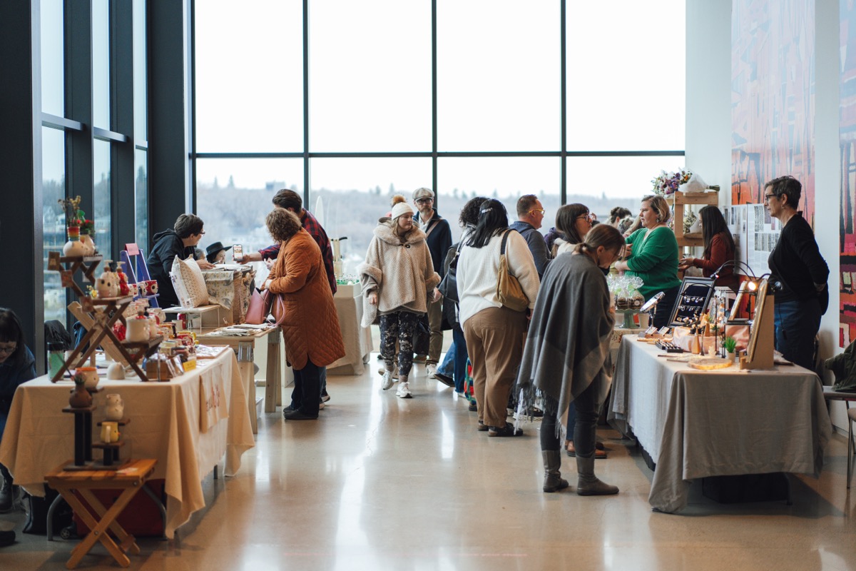 A bustling indoor market with vendors and visitors interacting at tables displaying handmade goods, art, and crafts. The space has large floor-to-ceiling windows letting in natural light and offering a view of trees and the river outside.
