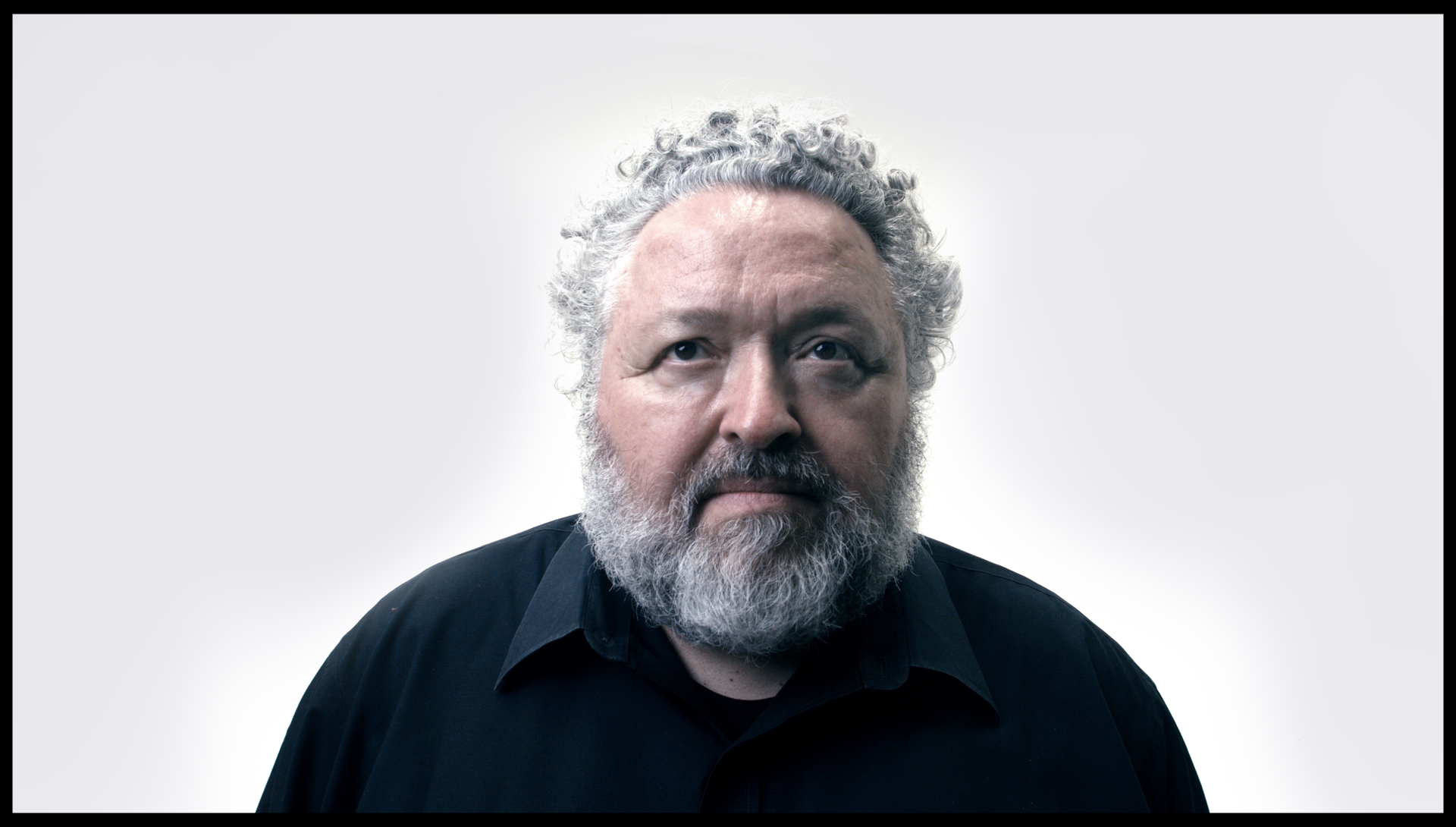 Portrait of a man with curly grey hair and a full beard, wearing a black collared shirt. He gazes slightly upward with a thoughtful expression. The background is a seamless, pale gradient, giving the image a clean and minimalist look.