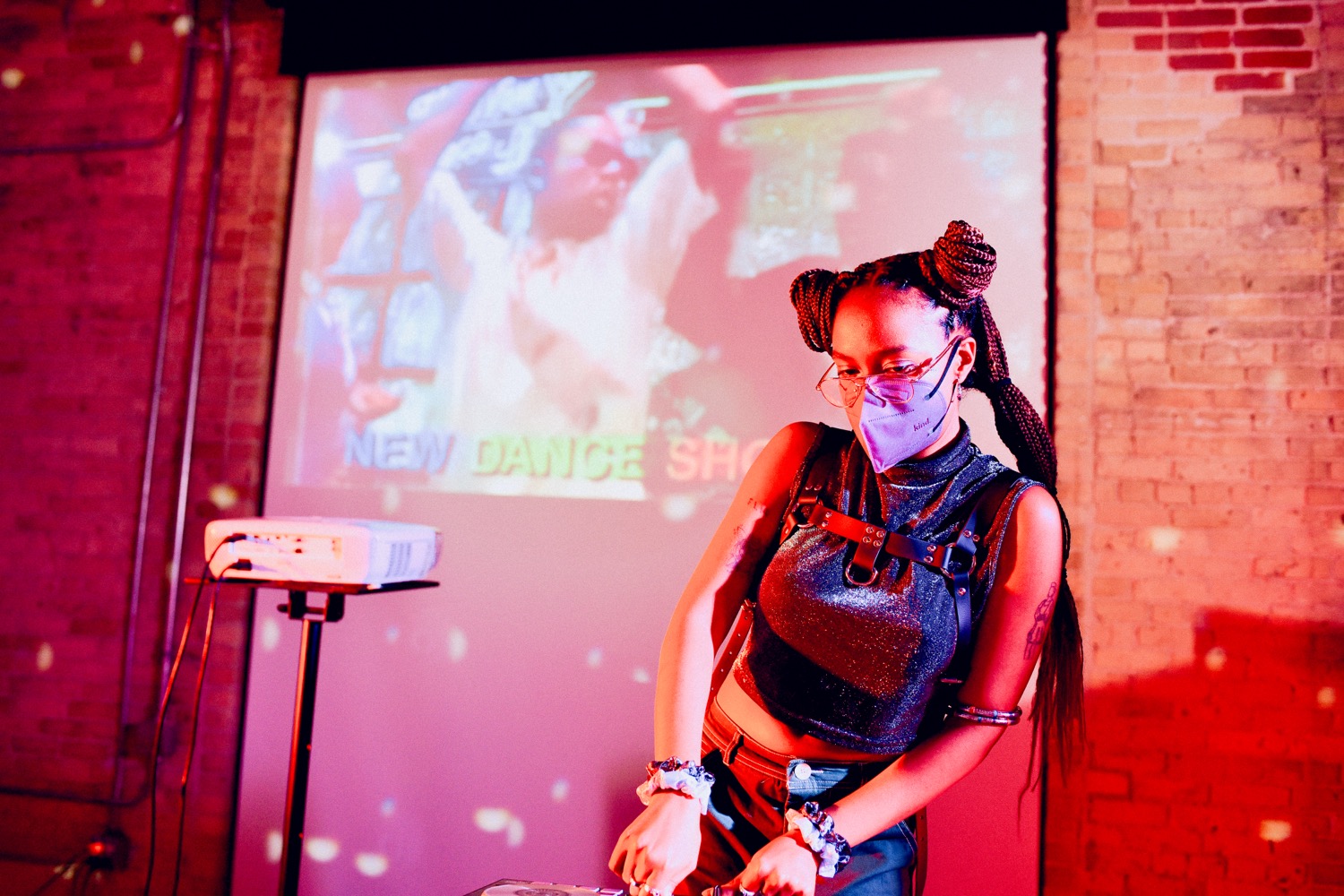 Mahlet Cuff performs as HYPERFEMME GALACTICA in a dimly lit space with pink and red lighting. They stand behind DJ equipment wearing a sparkly top, leather harness, and purple face mask, with a projected video and the words “NEW DANCE SHOW” visible on the wall behind them.