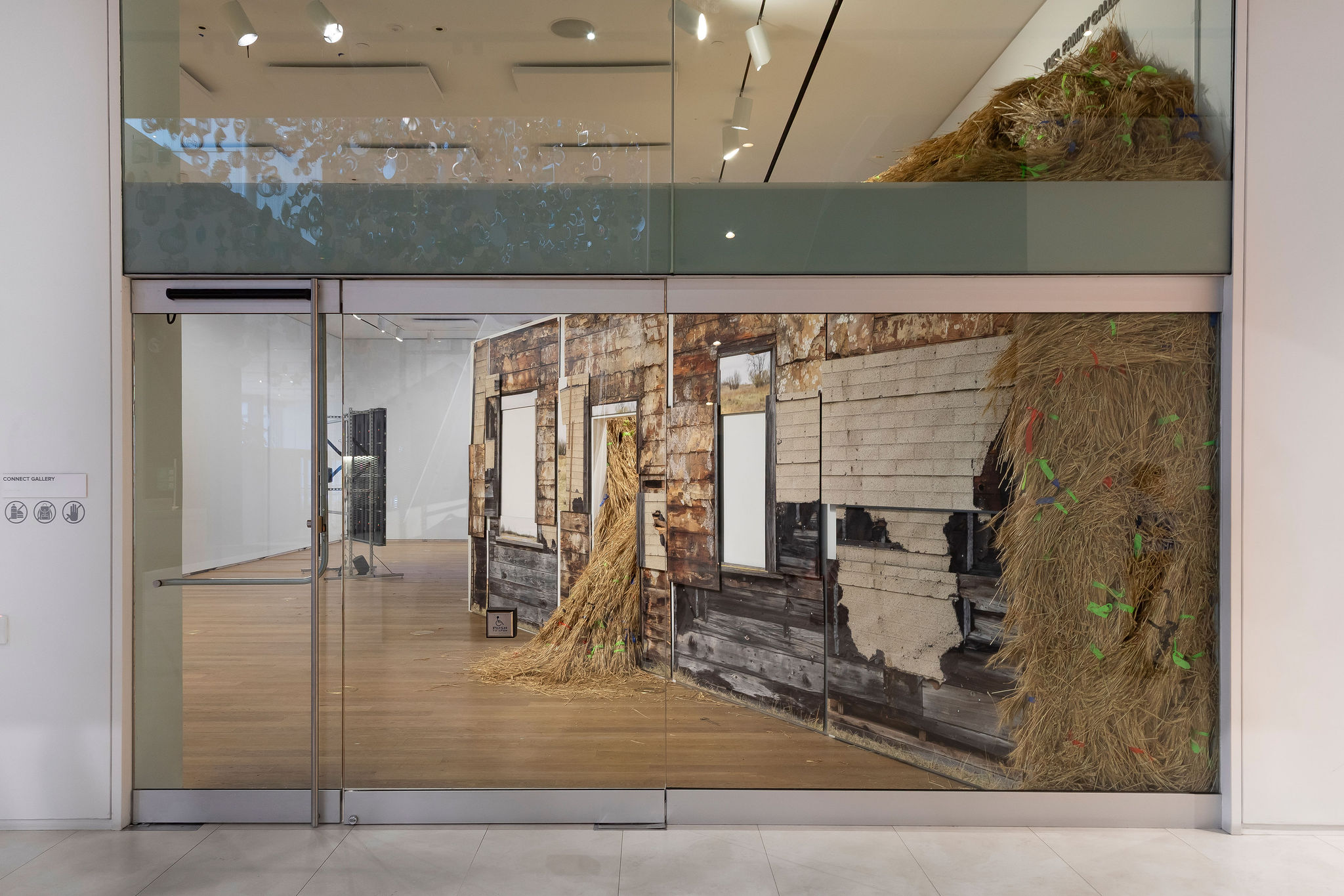 View through glass gallery doors of an installation resembling the façade of a weathered wooden house, with sections of straw spilling out from window openings and along the right side; bits of colorful ribbon are scattered throughout the hay, and the museum’s wood flooring and white walls frame the scene.