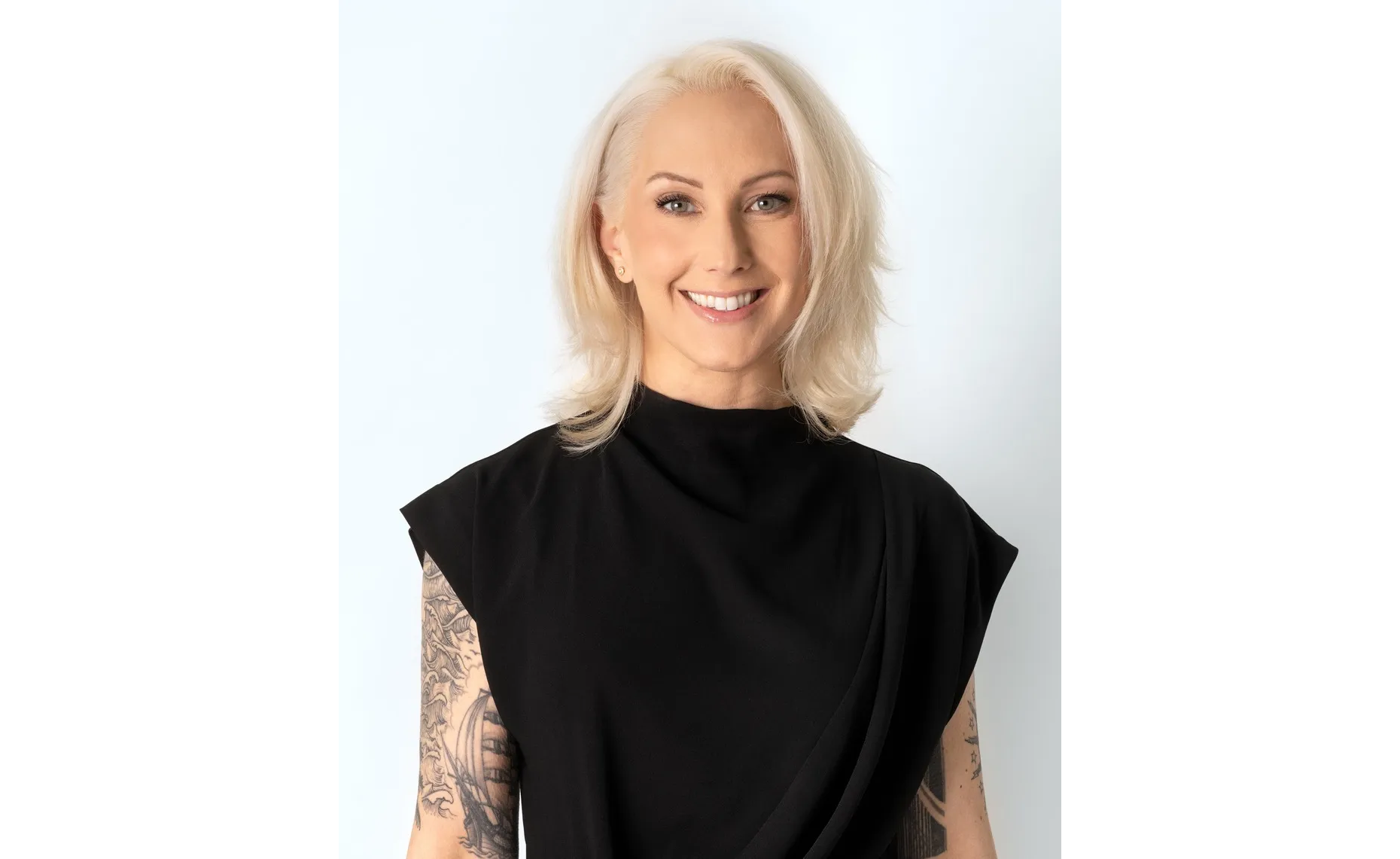 A smiling person with short, light-blonde hair wearing a sleeveless black top. They have visible tattoos on both arms and are standing against a plain, light background.