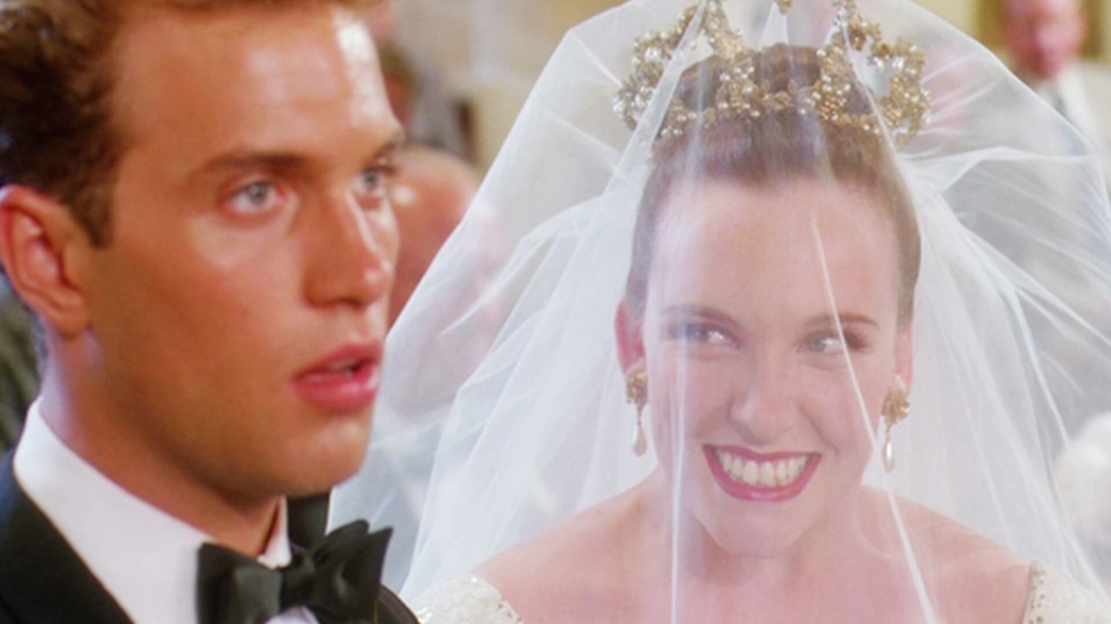 Close-up of a bride and groom at their wedding ceremony. The bride wears a sparkling tiara and white veil, beaming with an exaggerated smile. The groom, dressed in a tuxedo, looks forward with a stunned, dazed expression. The scene has a saturated, slightly dreamy quality.