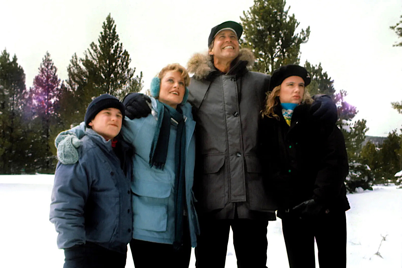 A family of four stands close together in a snowy forest, bundled in winter coats, hats, and scarves. They smile and gaze upward, with arms wrapped around each other. The sky is overcast, and tall pine trees frame the background, evoking a festive and heartwarming holiday atmosphere.