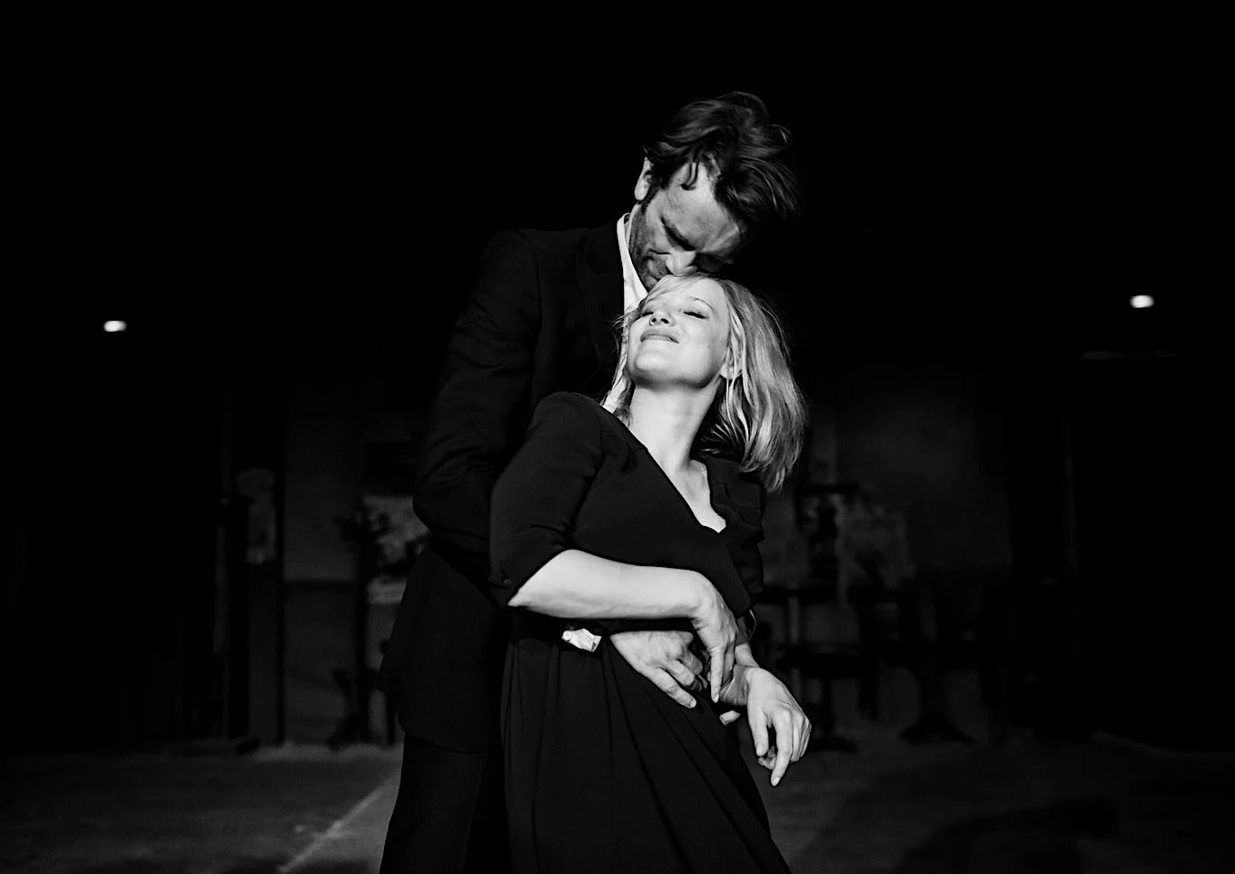 A black-and-white image of a man embracing a woman from behind, both dressed in dark clothing. The man gently presses his face to her head, eyes closed, while the woman leans back into him with a serene smile and closed eyes, illuminated against a dimly lit background.