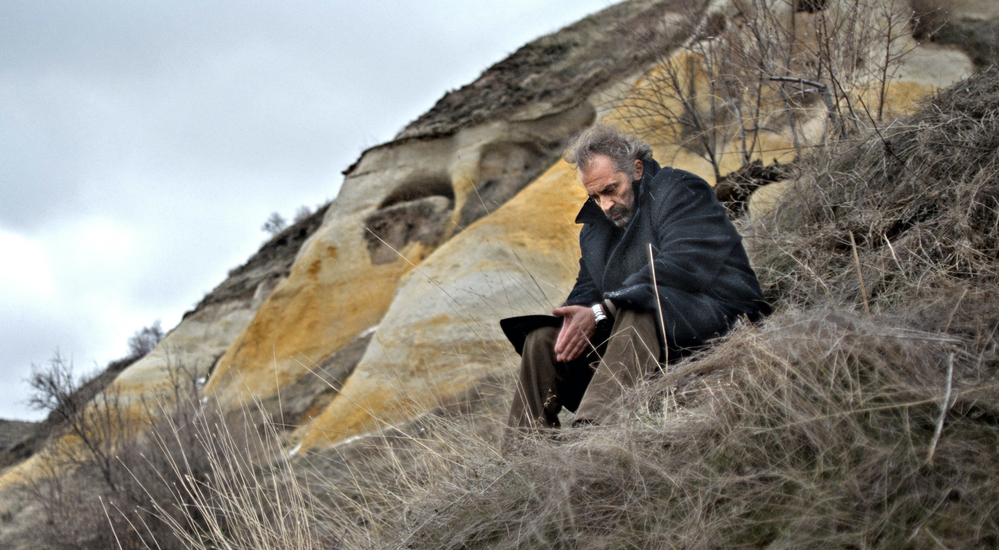 A man with greying hair and a heavy coat sits alone on a dry, grassy hillside, hunched forward in a contemplative pose. Behind him rise wind-carved, ochre-and-white rock formations under a cloudy sky. The scene evokes a sense of solitude and introspection amid a stark, natural landscape.