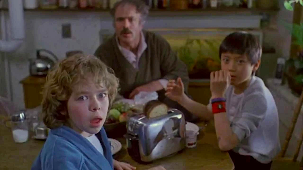 Three people sit around a kitchen table and look toward the camera with startled expressions. In the foreground, a child with curly hair and a blue robe has their mouth slightly open. Beside them, another child in a light shirt rests a hand near their face. An adult sits behind them at the table. A toaster, plates, bread, and food are spread across the table, with shelves and kitchen items visible in the background.