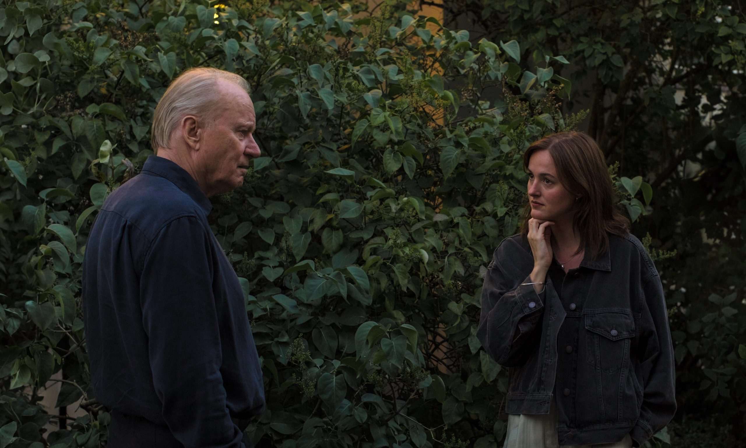 Two people stand facing each other in a quiet garden or courtyard dense with green foliage. An older man with thinning gray hair, dressed in a dark shirt, looks downward with a somber, reflective expression. Across from him, a younger woman in a dark jacket rests her hand against her chin, studying him thoughtfully.