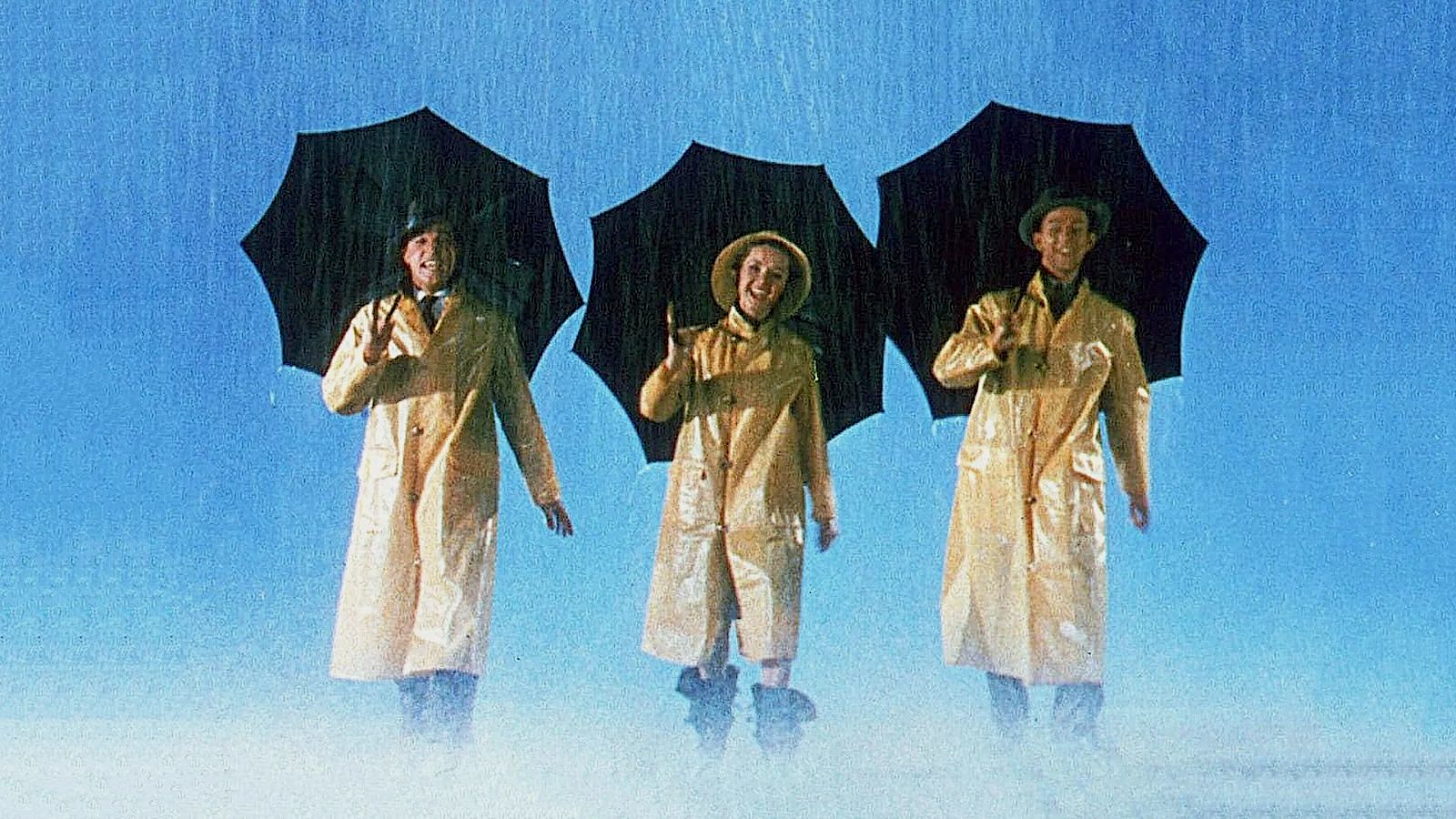 Three people walk side by side through heavy rain, each holding a black umbrella. They wear matching light-coloured raincoats and hats, smiling and gesturing as they move forward. The background is a bright blue sky, and rain splashes around their feet.
