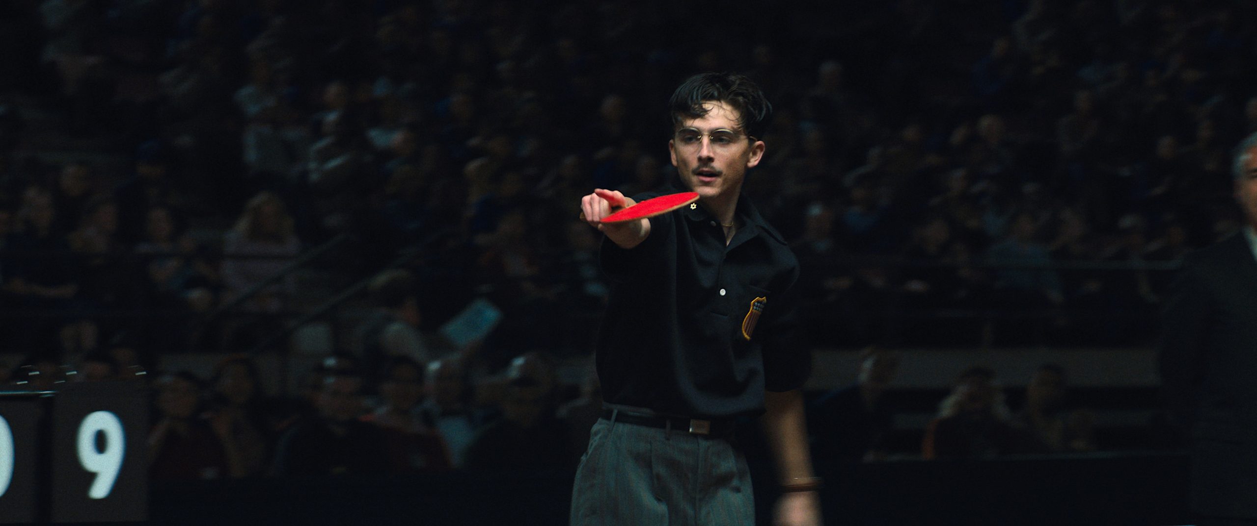 A young man stands inside a darkened arena or sports hall, extending his arm forward while holding a bright red table tennis paddle. He wears a black shirt and gray trousers.
