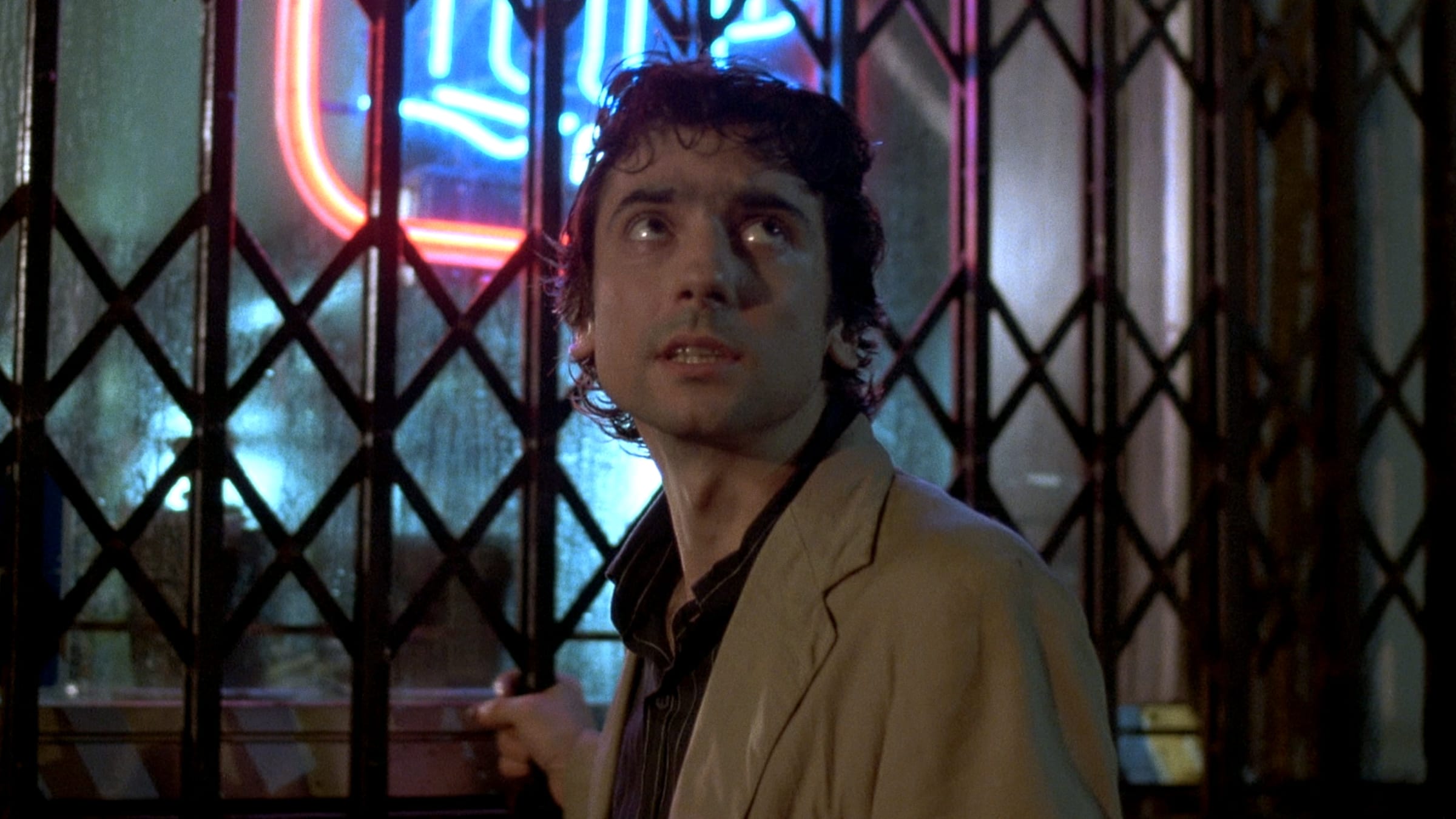 A man stands behind a metal security gate at night, looking upward with a tense, uncertain expression. He wears a light-colored jacket over a dark shirt. Behind him, a window glows with blue and red neon light, casting reflections on the glass and emphasizing the nighttime, urban setting.