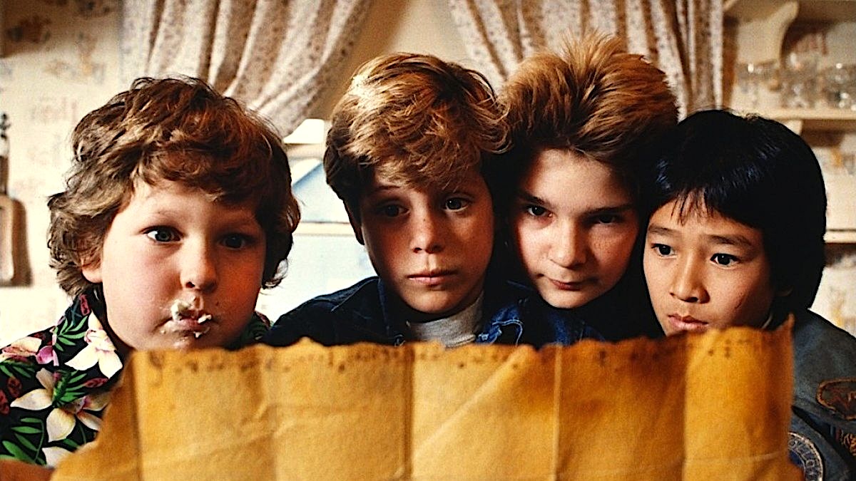 Four children lean closely together indoors, peering intently at a large, aged paper held in front of them. One child has food smeared around their mouth, while the others look focused and curious. Warm indoor lighting, patterned curtains, and shelves in the background create a cozy, adventurous atmosphere.