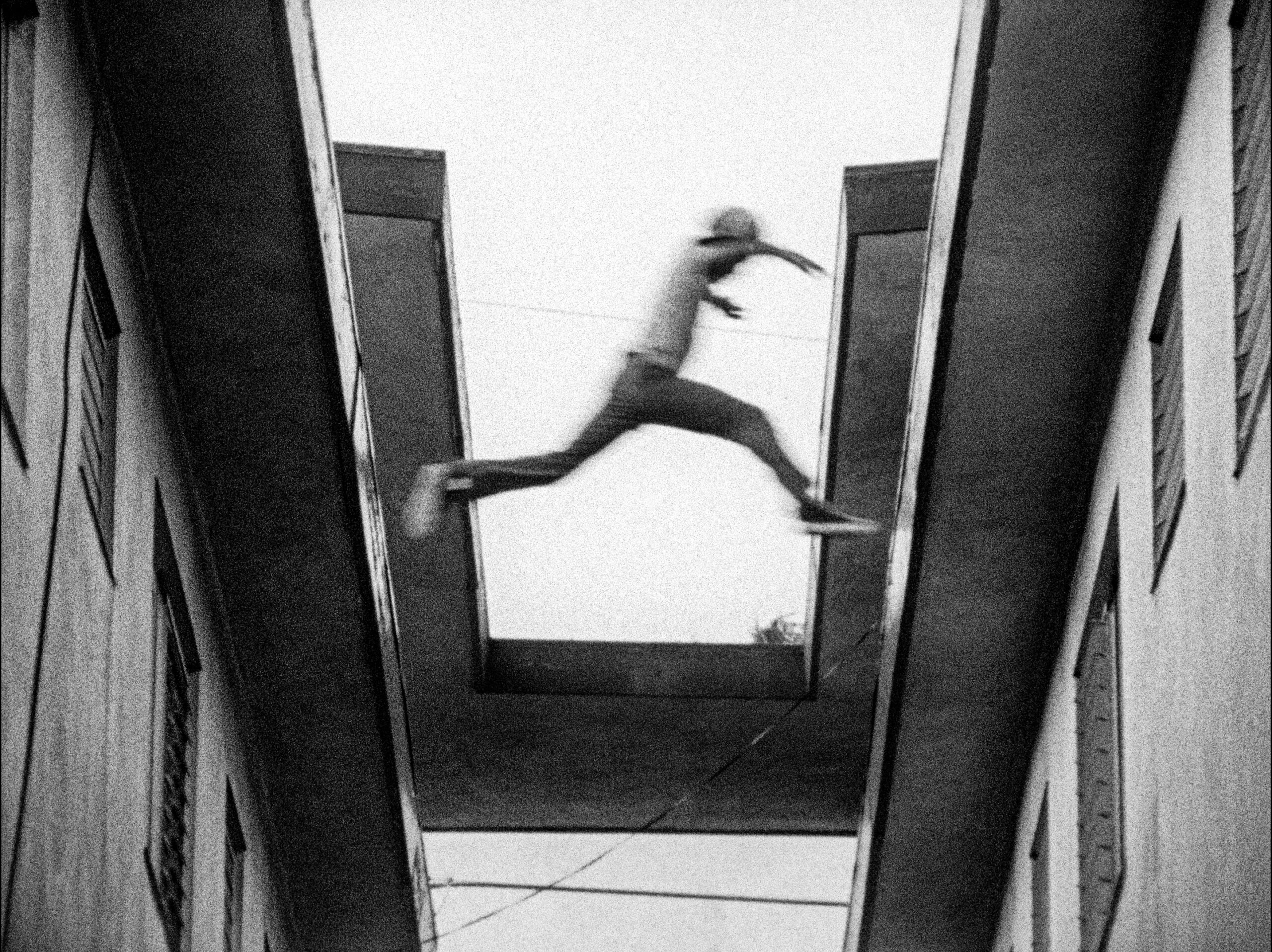 A black-and-white image shows a person mid-jump, leaping across a narrow gap between two concrete buildings. The figure is blurred in motion, legs stretched wide as they cross the open space below. Stark architectural lines and deep shadows frame the scene.