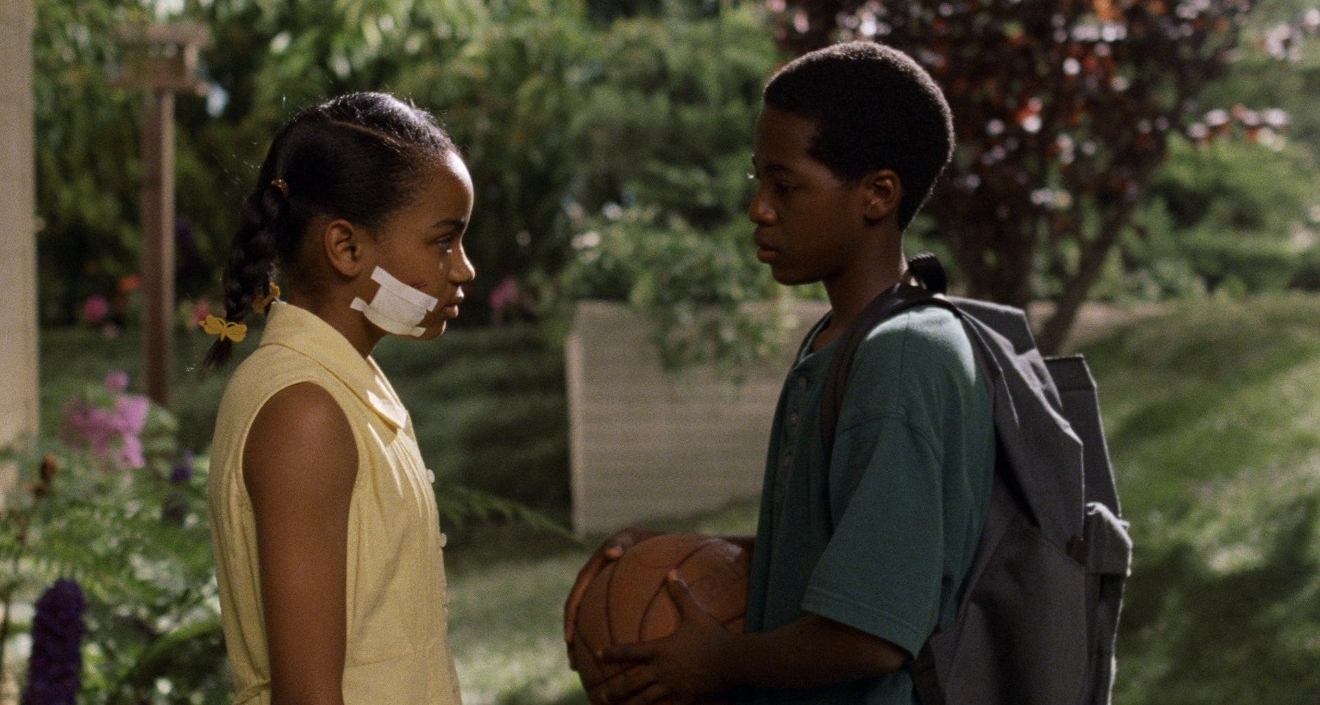 Two children stand facing each other outdoors in a leafy residential setting. One child wears a yellow dress and has a bandage taped to their cheek, while the other wears a green shirt and a backpack and holds a basketball.
