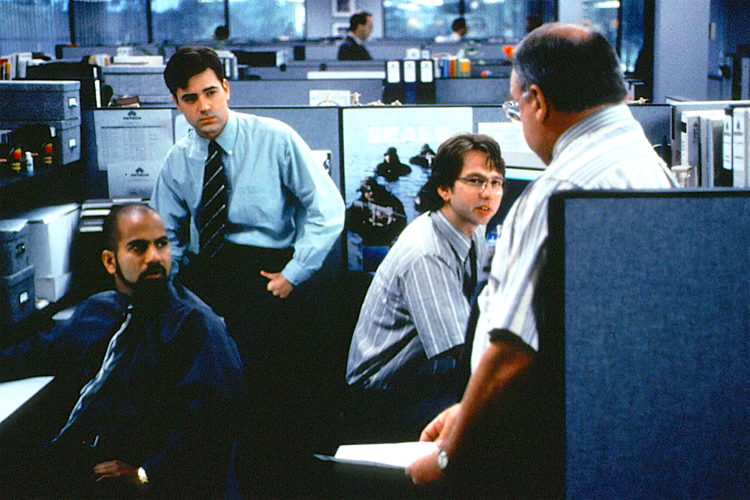 Several men gather around cubicles in a busy office with gray partitions and computer monitors. One man sits at a desk while others stand nearby, listening to an older man who appears to be addressing them. They wear office attire such as button-down shirts and ties. The background shows more cubicles and coworkers.