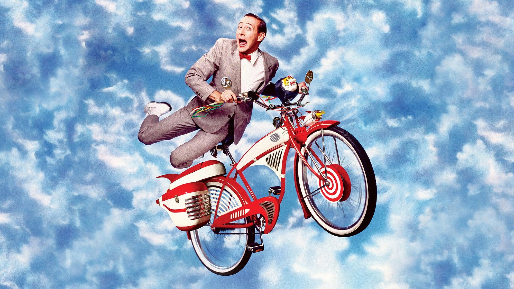 A man in a gray suit with a red bow tie rides a red-and-white bicycle high in the air, as if flying against a bright blue sky filled with fluffy clouds. He grips the handlebars with one hand and kicks a leg out behind him, wearing an excited, wide-eyed expression.
