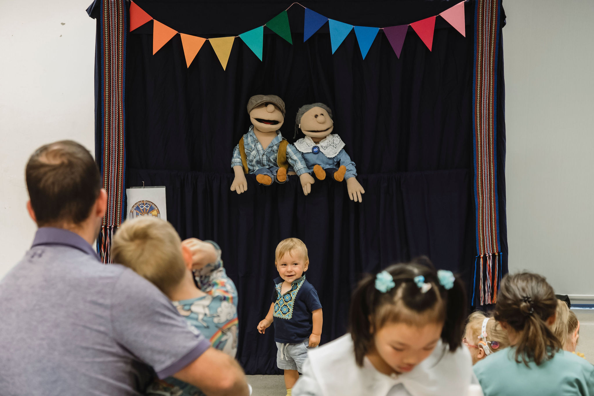 Children and caregivers gather in front of a small puppet theatre decorated with colourful triangular bunting. Two fabric puppets sit on a dark curtained stage, while several young children stand and move about in the foreground, including a smiling toddler at centre facing the camera.