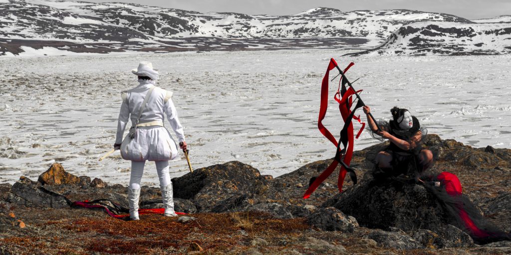 Two figures stand on dark rocks at the edge of a frozen or icy body of water, with snow-covered hills in the background. One figure, dressed entirely in white, faces the landscape, while the other, dressed in black and red with a face painted white, raises a stick adorned with long red ribbons.