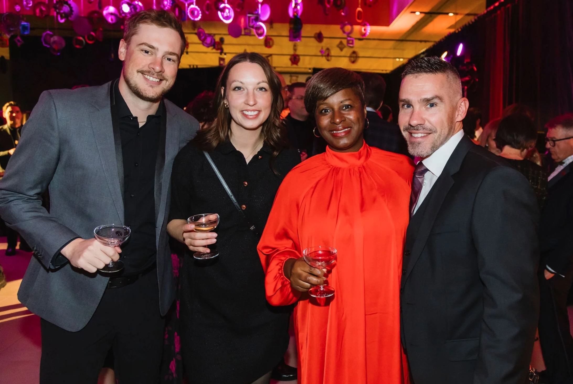 Four adults dressed in formal evening wear stand together at a gallery event, smiling and holding drinks. The space is warmly lit with pink and purple lighting, with decorative elements hanging overhead and other guests visible in the background.