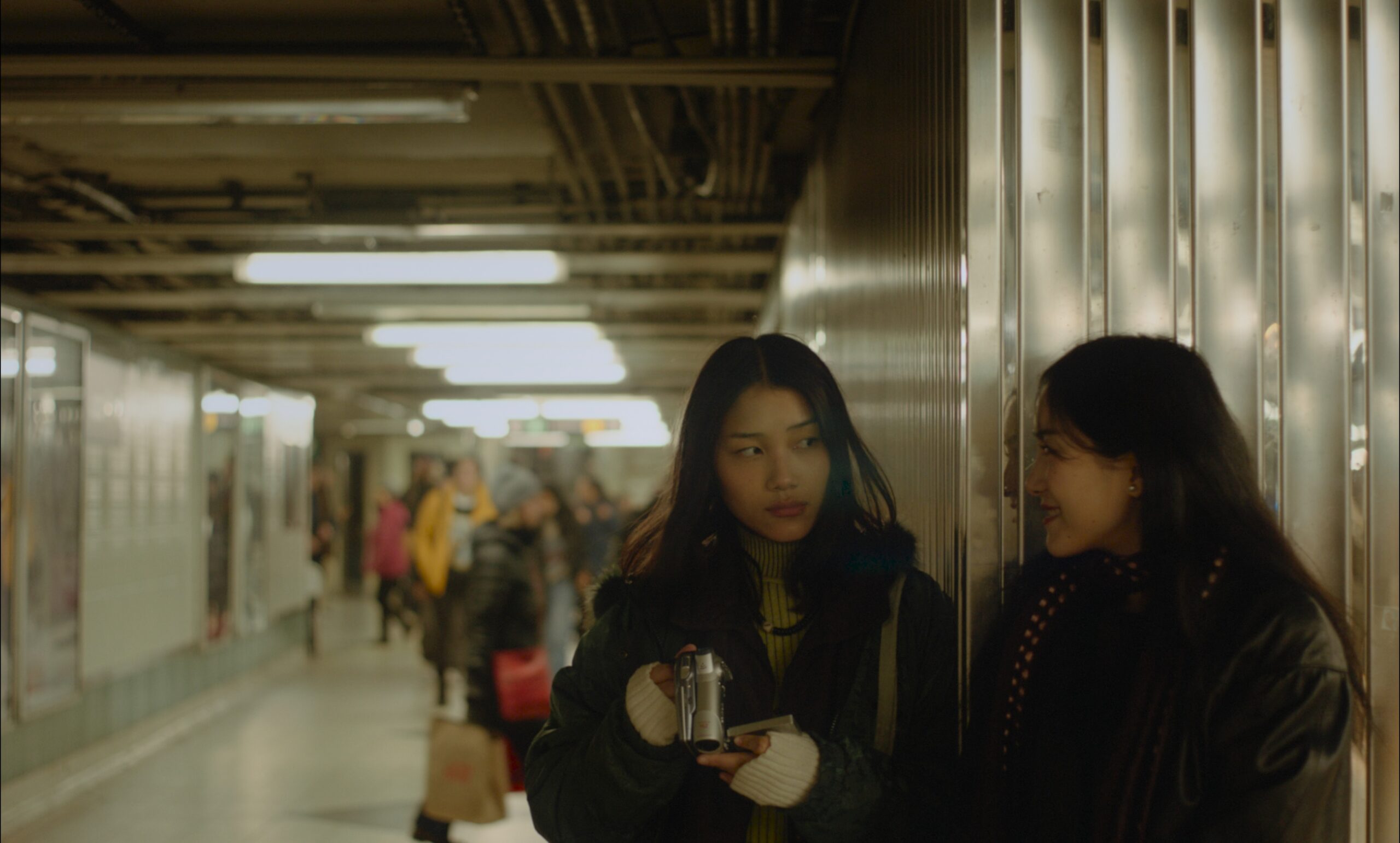 Two young women stand against a ribbed metal wall in a busy underground subway corridor, with blurred commuters moving through the tunnel behind them. One holds a small compact camera and gazes forward with a neutral expression, while the other leans against the wall smiling softly at her, both wearing dark winter jackets.