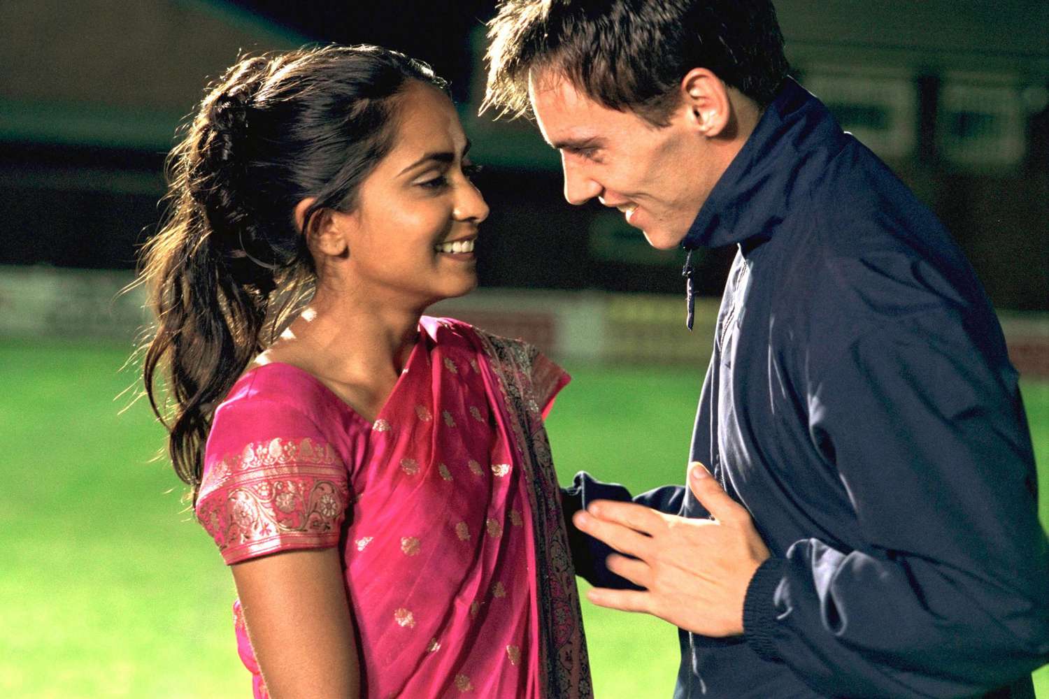 A young woman in a pink embroidered sari and a man in a dark zip-up jacket face each other closely on what appears to be a sports field at night, both smiling warmly.