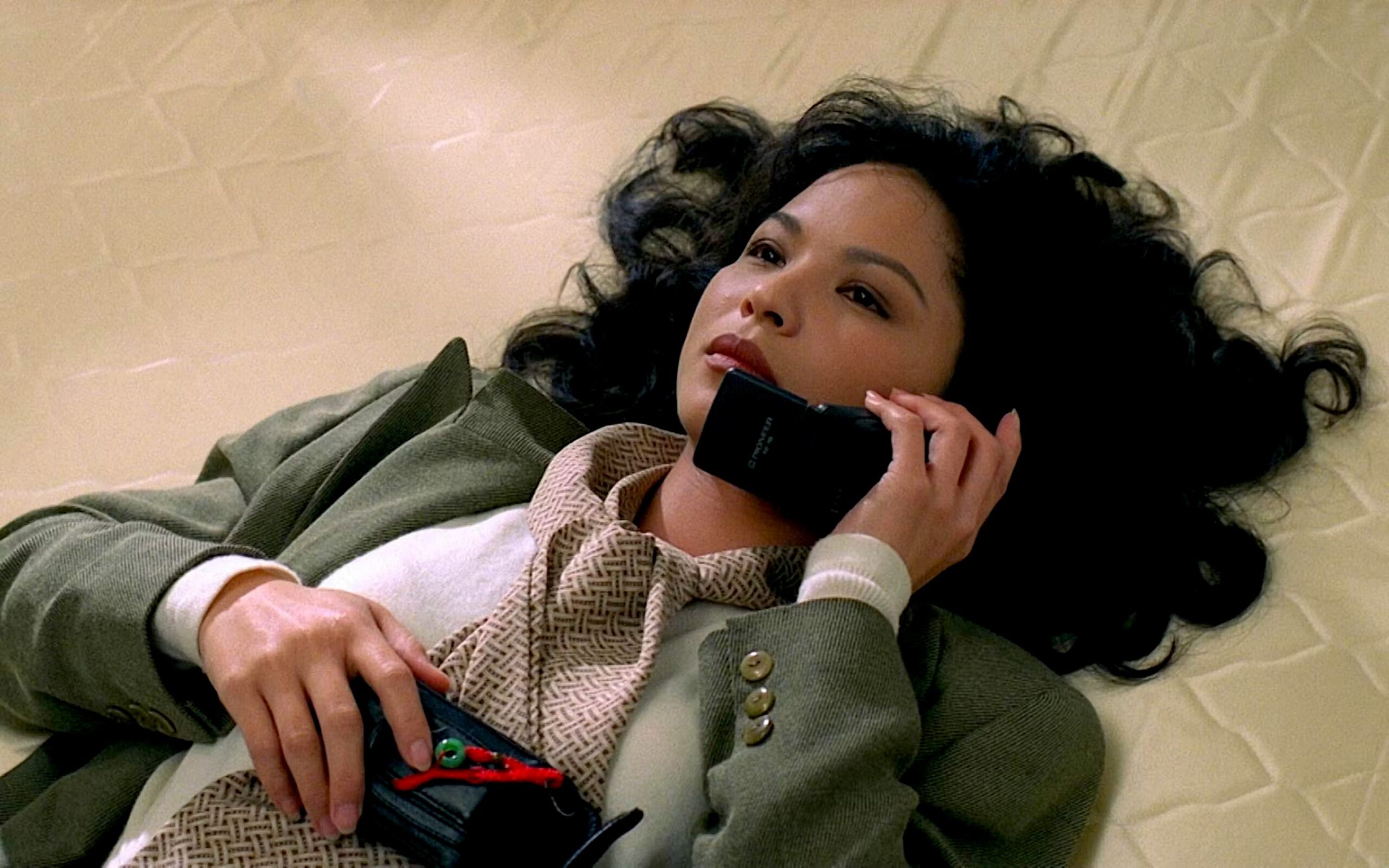 A woman with voluminous dark curly hair lies on a tiled floor, holding a vintage mobile phone to her ear while clutching a small bag to her chest. She wears a green blazer over a patterned scarf and cream sweater, gazing upward with a calm, contemplative expression. The film grain, styling, and chunky early-era cell phone suggest a movie still from the 1990s.