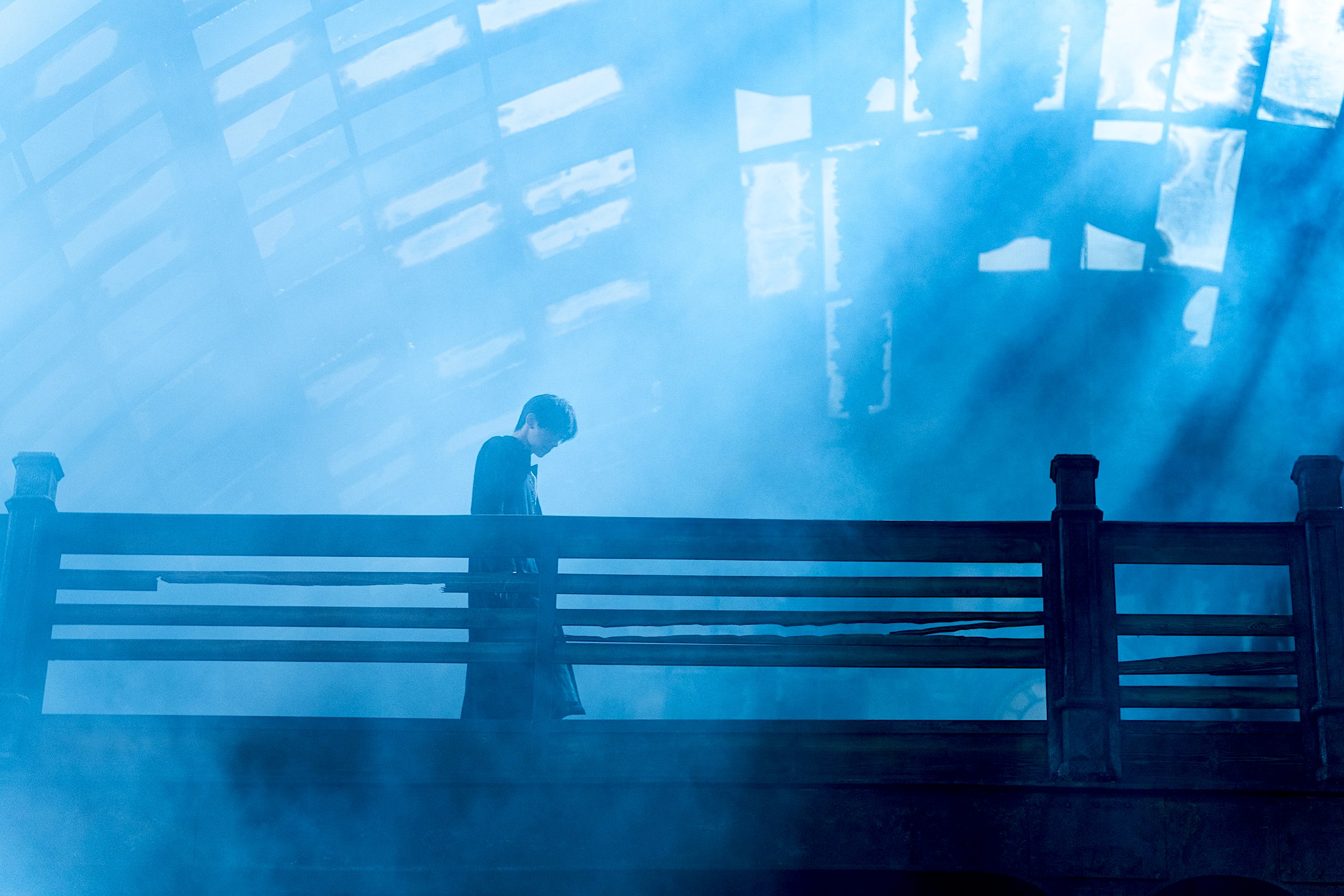 A solitary figure stands behind a railing, head bowed. Dramatic rays of light cut diagonally through the haze from a gridded glass ceiling above, creating an atmospheric, cinematic scene.
