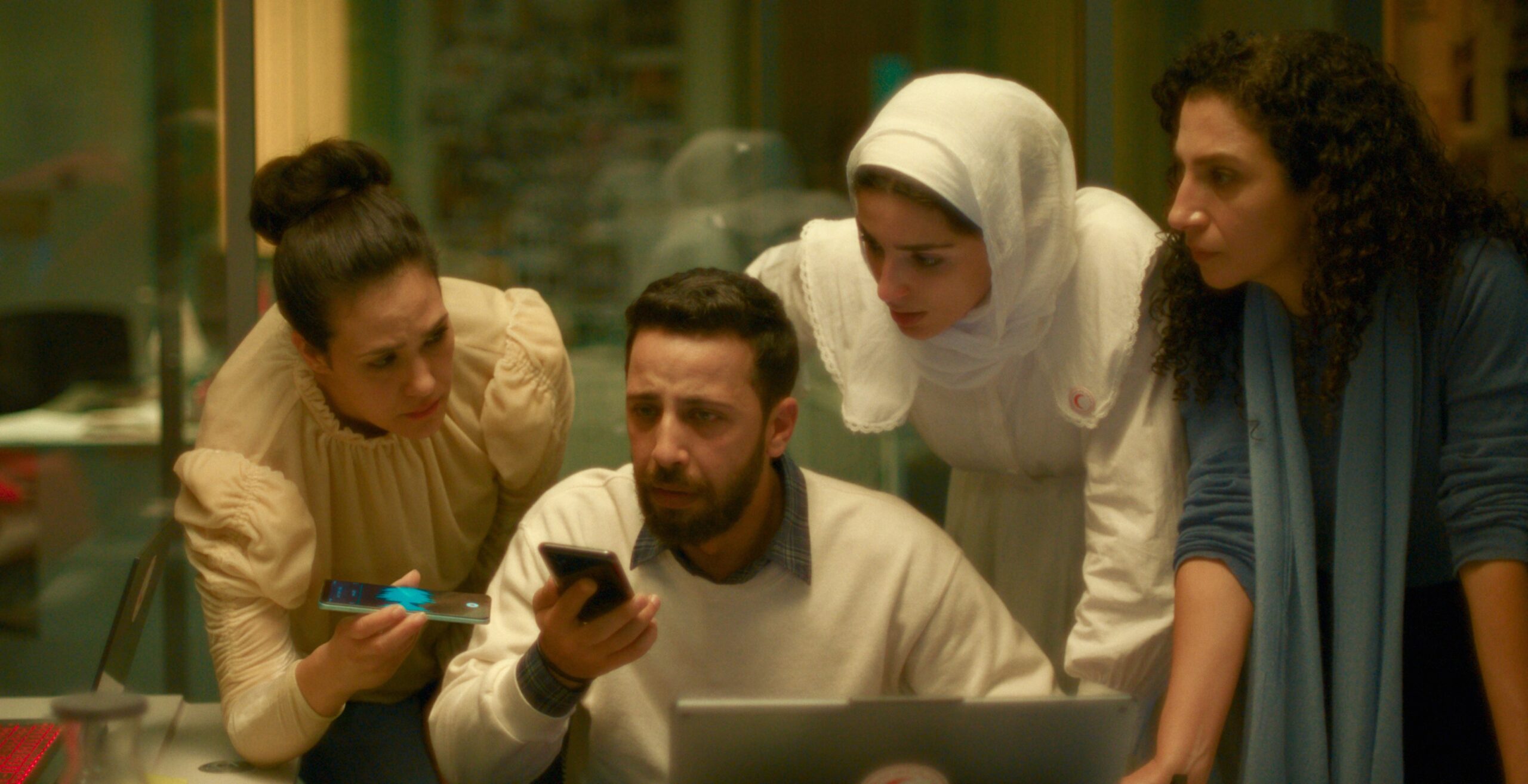 Four people huddle together around a laptop screen with serious, focused expressions. A seated man in a white sweater holds a smartphone while three women lean in around him — one in a cream blouse with her hair up, one wearing a white hijab with a red crescent emblem on her sleeve, and one in a blue top with curly hair.