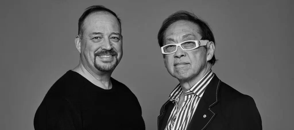 Black and white portrait of two smiling men standing side by side. One wears a dark sweater, the other wears a striped shirt with dark jacket and white-framed glasses.